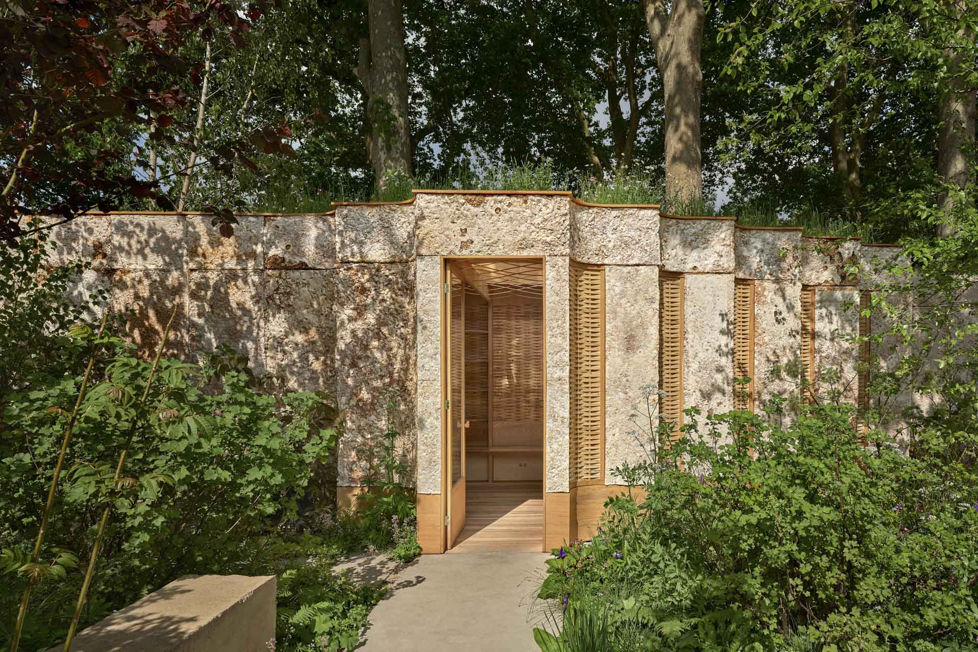 Kleines Haus im verwachsenen Umfeld, geschwungene Fassade, Flachdach begrünt, Holzsonnenschutz an Fenstern, offene Tür, Blick ins Innere.