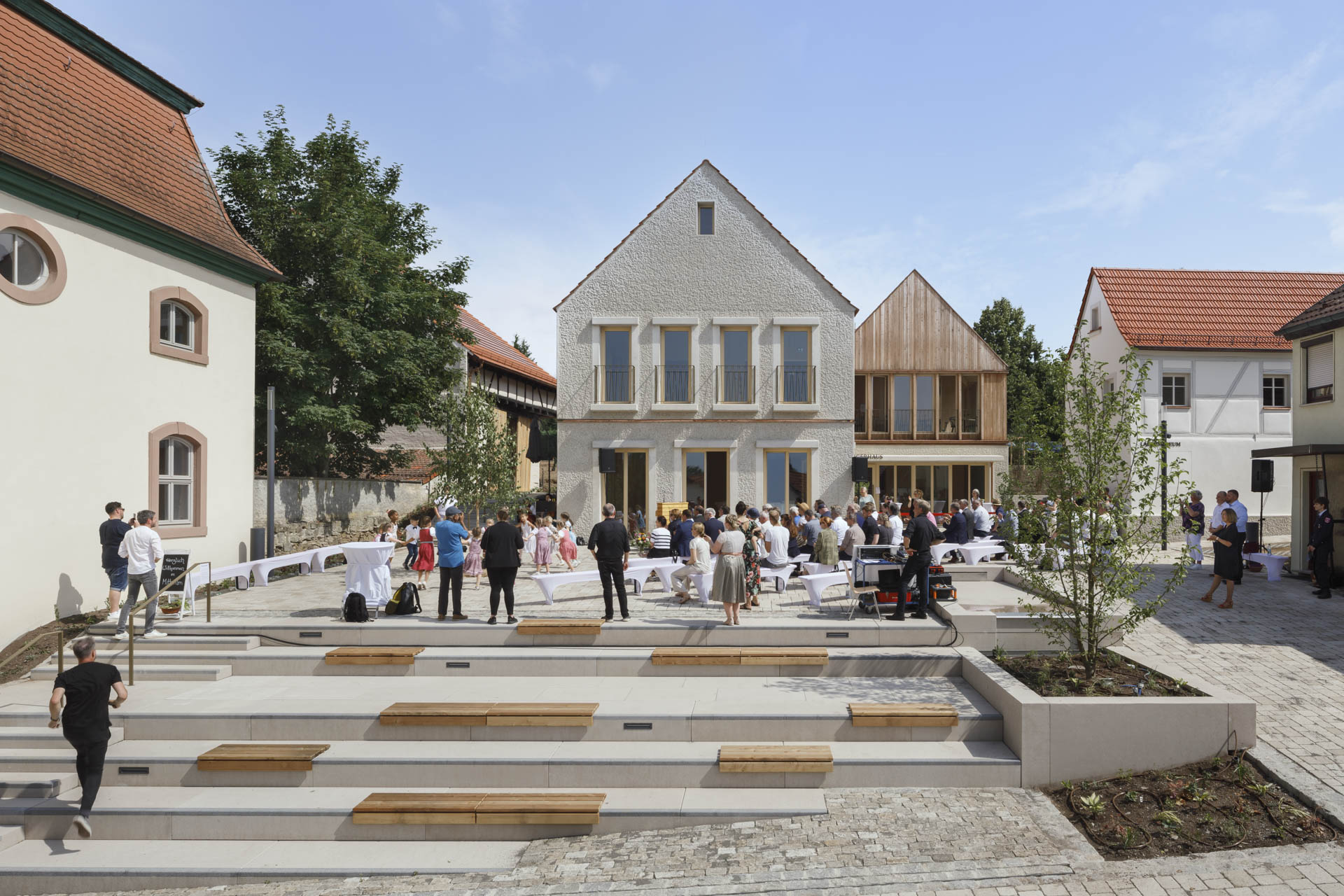 Dorfplatz mit vielen Menschen, gepflastertem Boden und flacher Sitztreppe.
