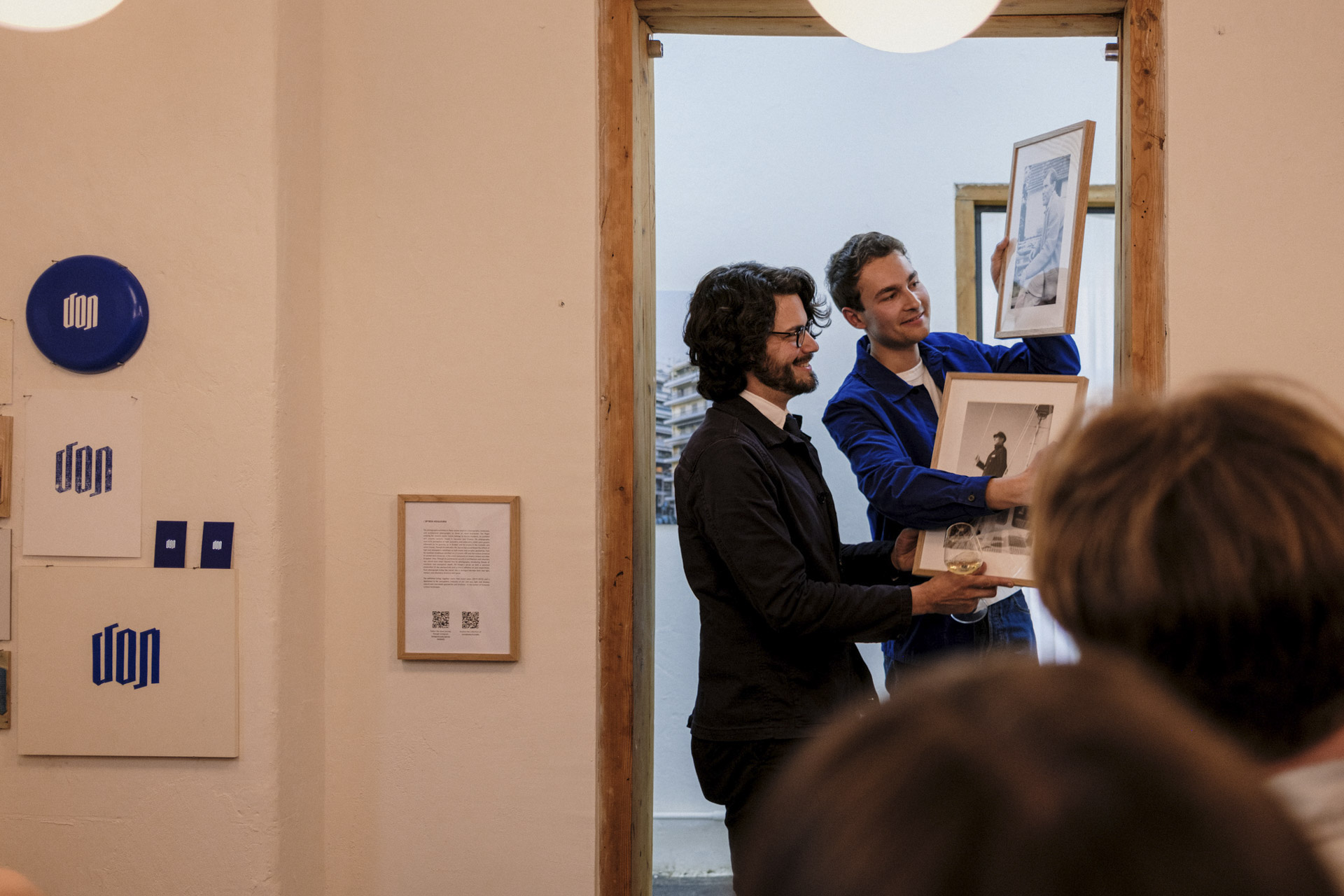 Zwei junge Herren stehen in einem Türrahmen einer Galerie und halten jeweils ein Bild in der Hand.