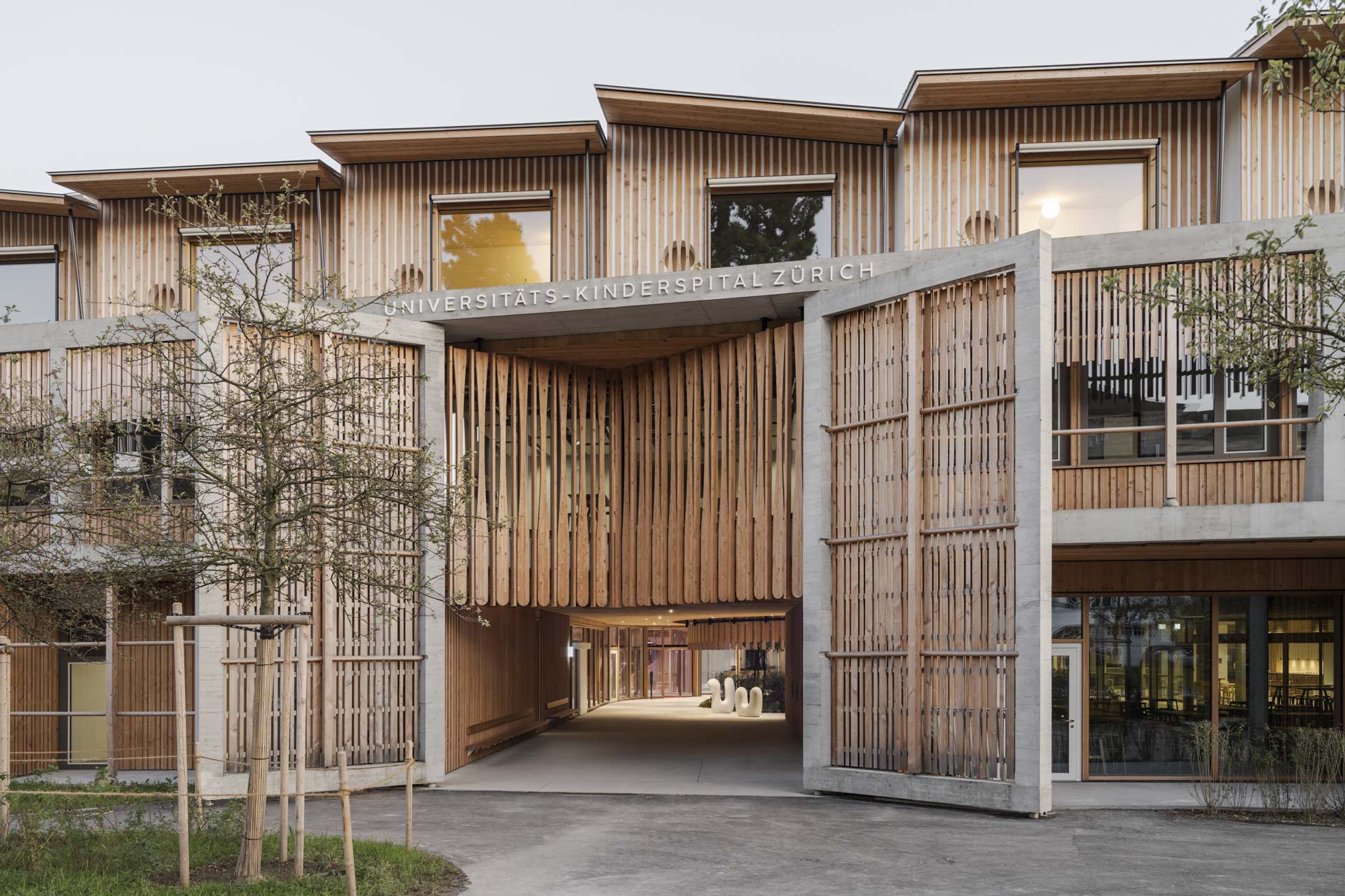 Eingangsbereich mit breitem Durchgang aus Holzlamellen und Betonrahmen, vertikale, rhythmische Struktur und großen Öffnungen.