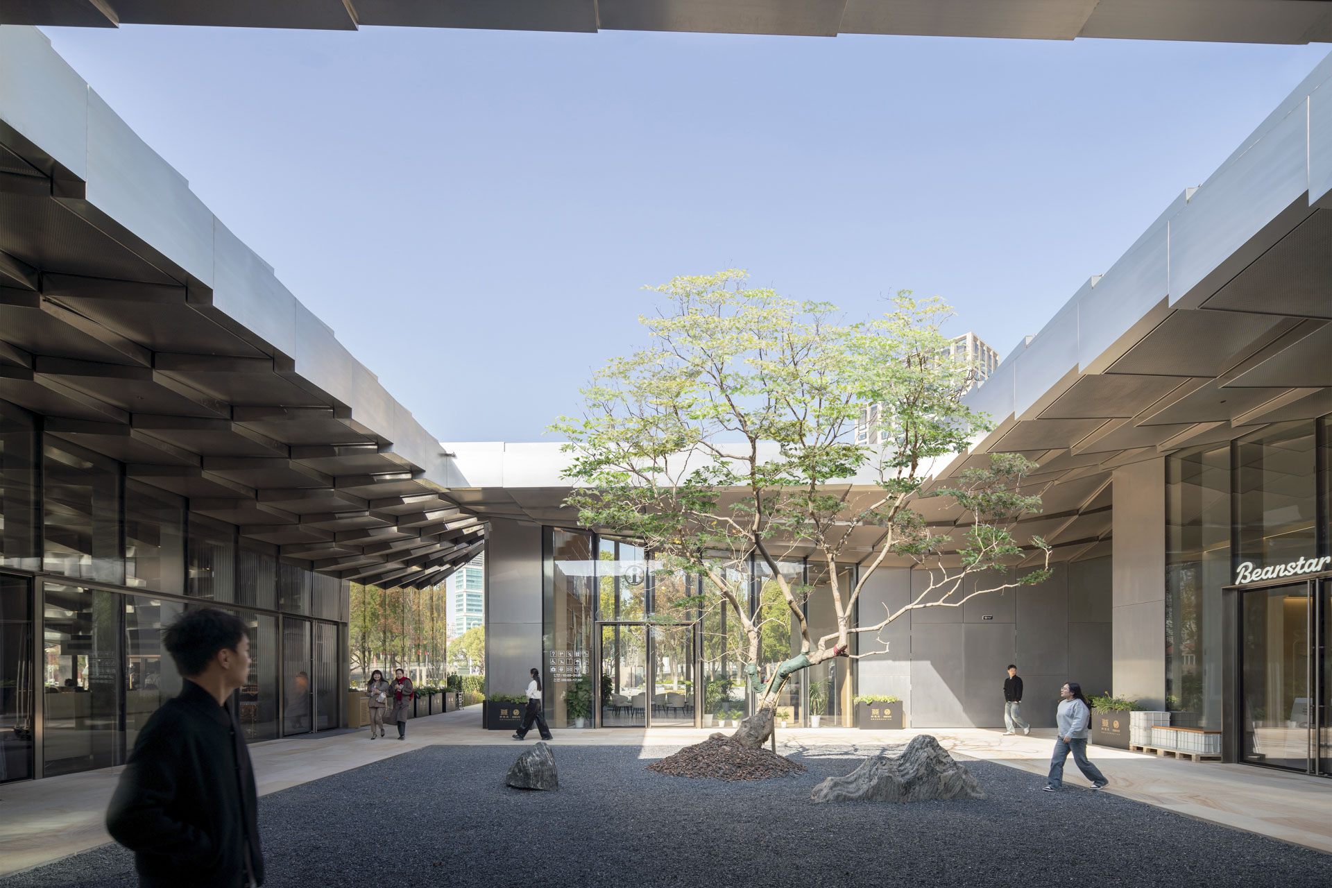 Modern courtyard with gravel floor, individual rocks and a tree in the middle. Surrounded by glass facades and people passing through the space.