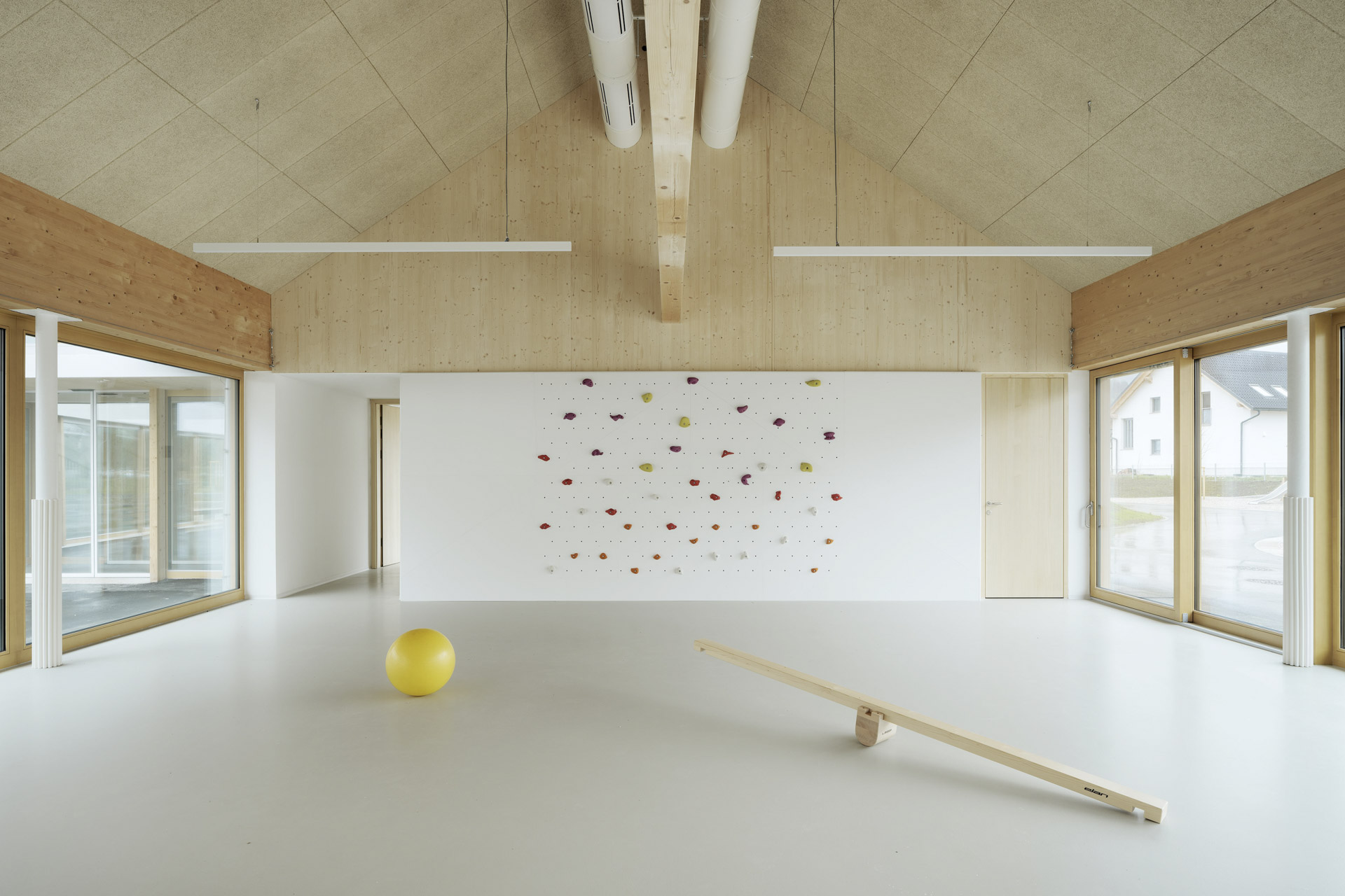 Innenraum unter Dachgiebel mit sichtbaren Holzelementen, Kletterwand, Wippe, großem Ball und großen Fenstern, heller Raum.