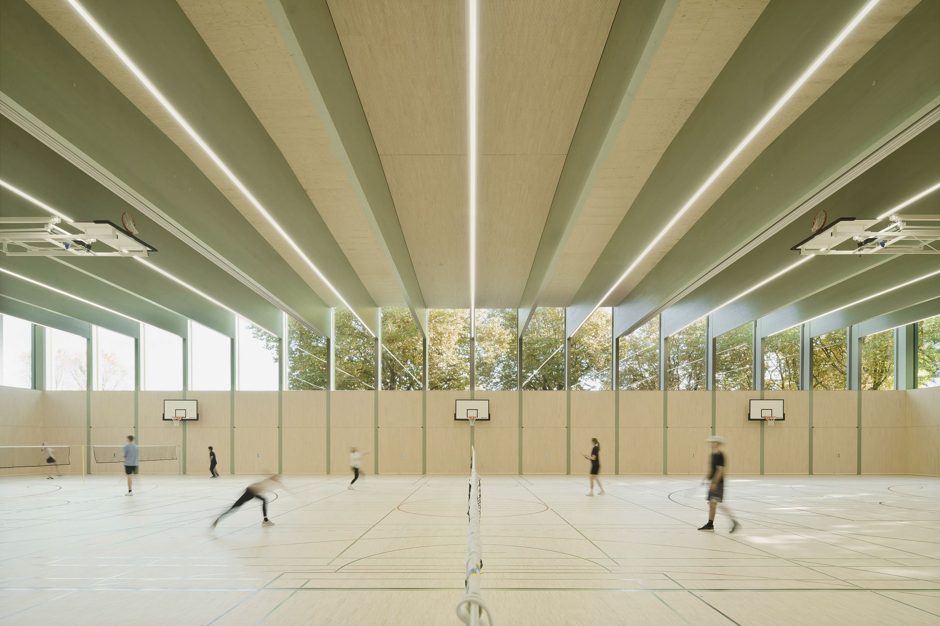 Light-filled sports hall with tall windows, wood paneling, and exposed ceiling beams, flexible playing area with basketball and badminton courts.