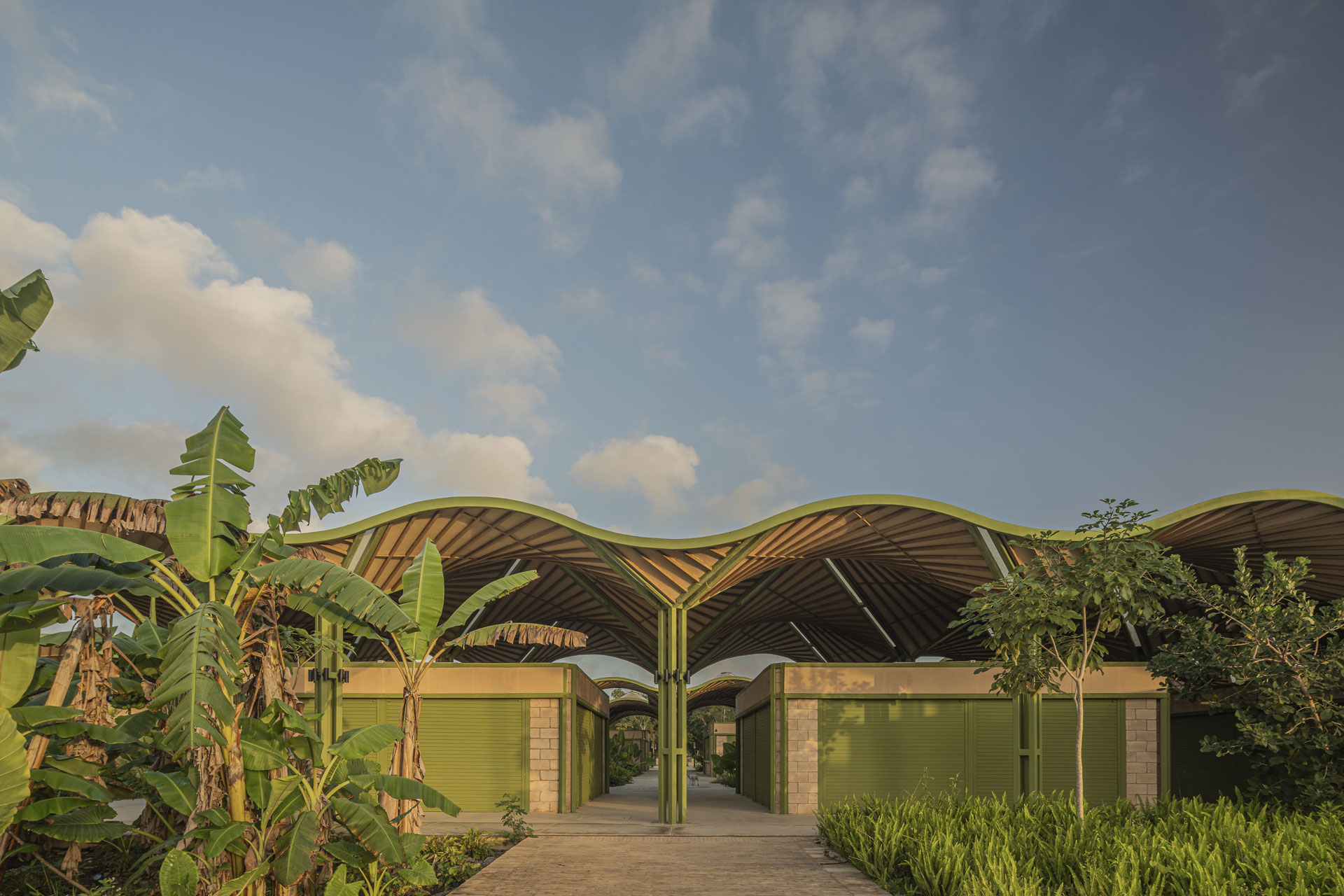 Gebäude mit gewelltem Dach aus Holzlamellen und grün eingefasster Struktur, flankiert von modularen Baukörpern mit grünen Rollläden und Vegetation.