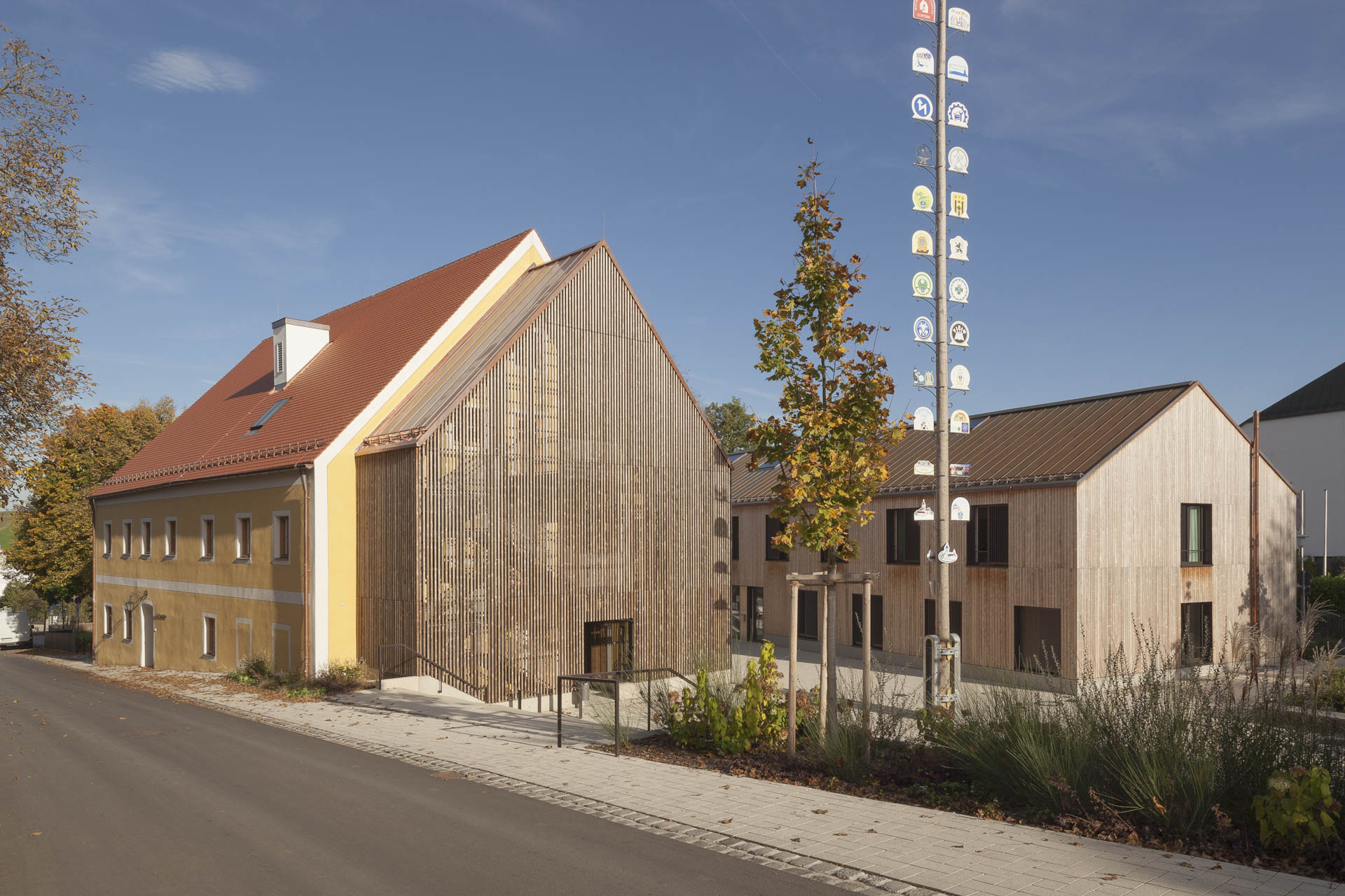 Traditionelles Gebäude mit gelber Fassade und rotem Satteldach trifft auf modernes Holzvolumen, umgeben von Grün und gepflastertem Vorplatz.