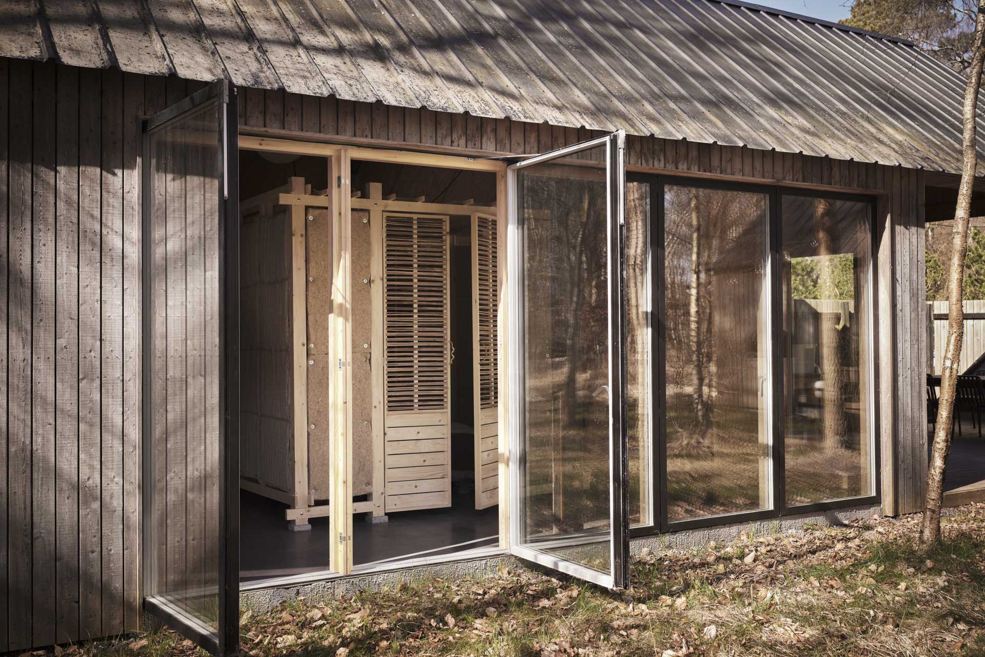 Wood-clad building with sloping metal roof, large folding glass doors, and exposed wood construction in the interior.