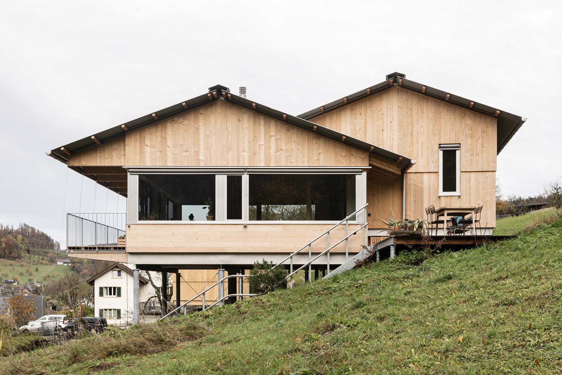 Haus in Hanglage mit heller Holzfassade, großem Fensterband, Metalltreppe und vorgelagerter Terrasse.