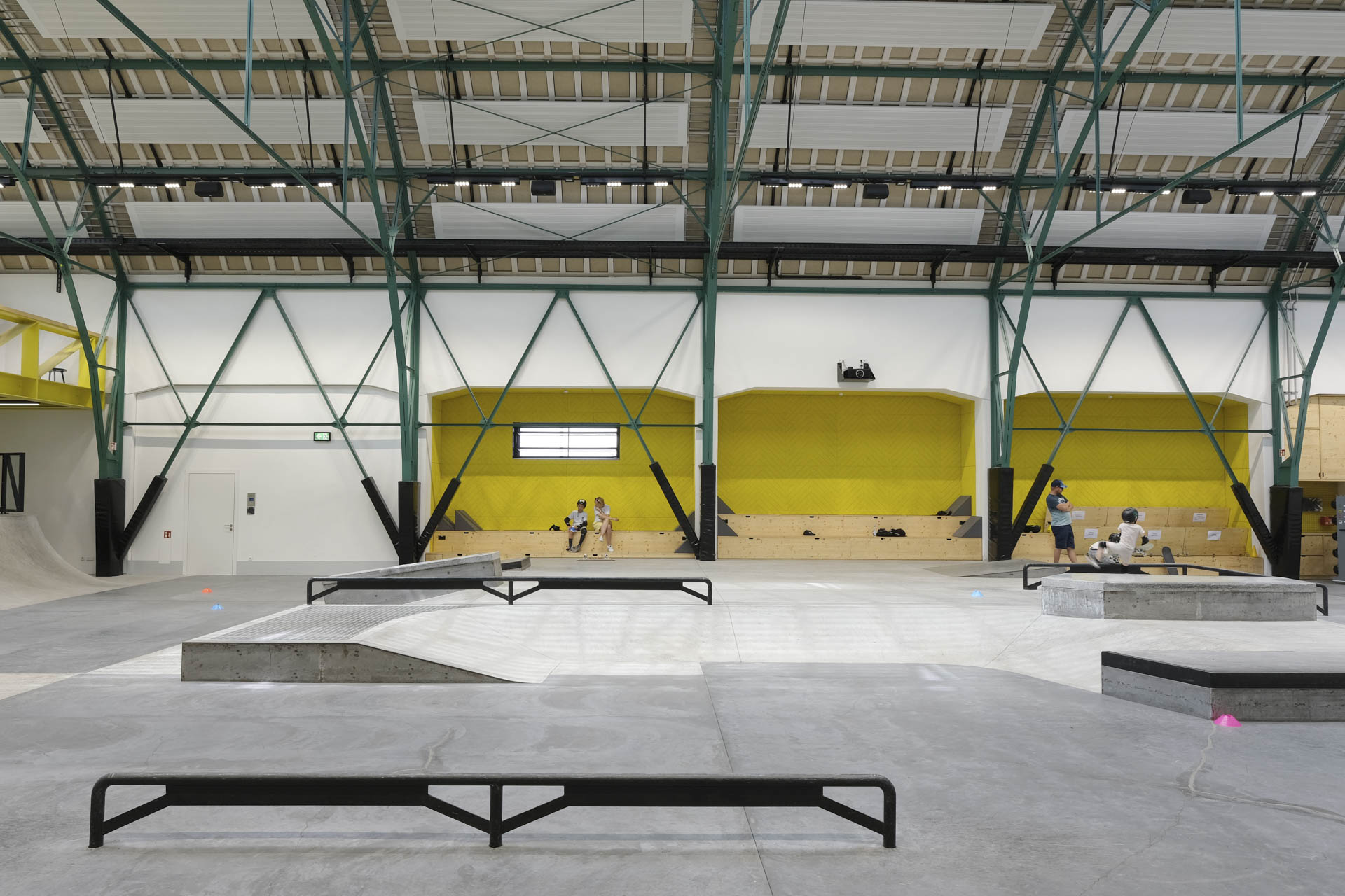 Innenraum mit betoniertem Skaterplatz, grüne Fachwerkträger tragen das Dach, gelbe Wandnischen mit Sitzbänken, Kinder skaten.
