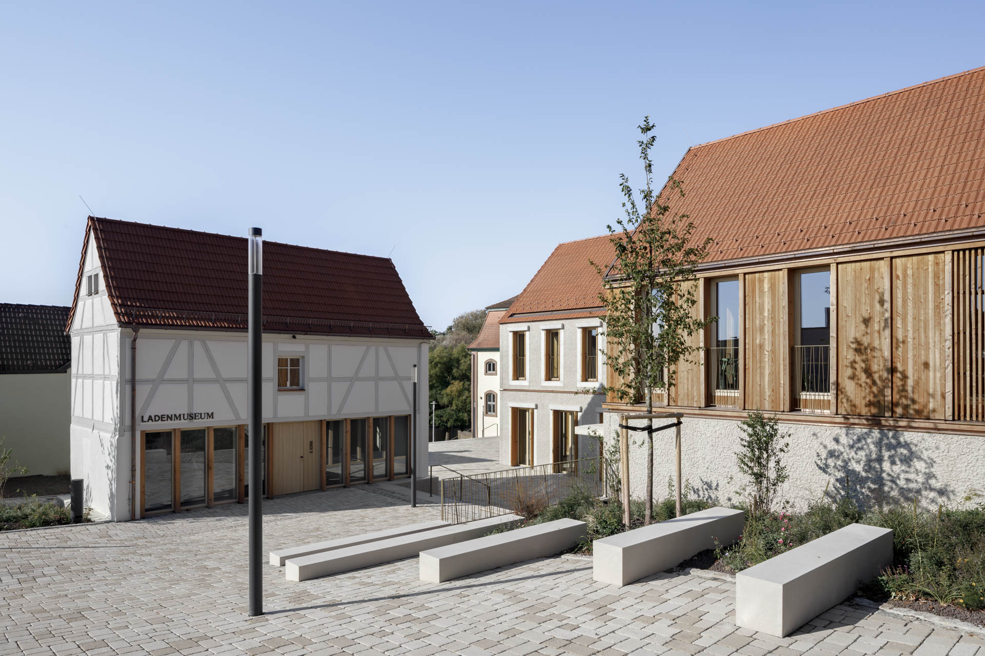 Gepflasterter Dorfplatz umgeben von niedrigen Gebäuden im ländlichen Stil. Häuser mit traditionellen Giebeldächern prägen das Bild.