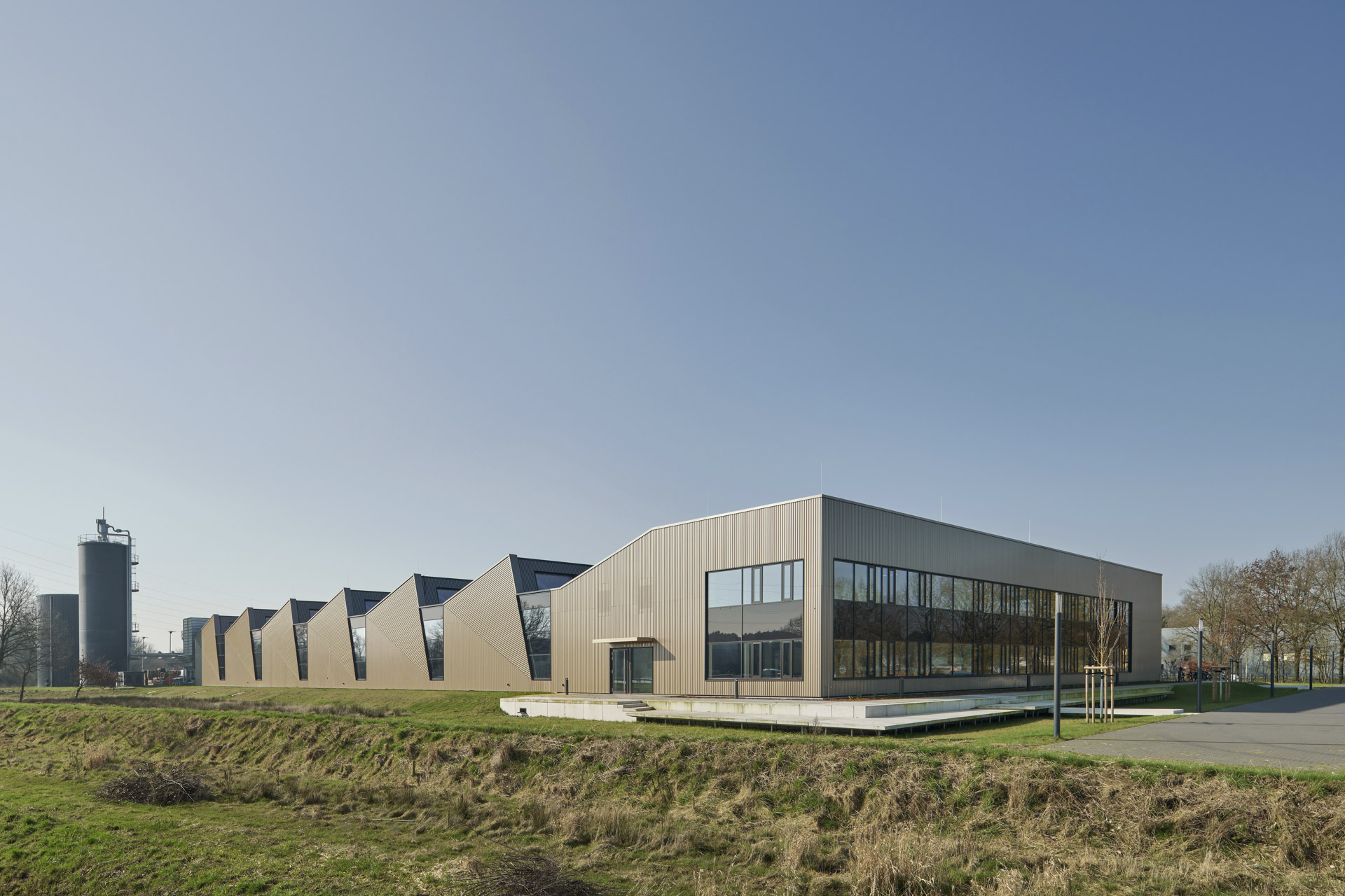 Außenansicht eines Industrie- oder Bürogebäudes mit gezacktem Sheddach, vertikaler Metallverkleidung und großflächigen Fensteröffnungen.