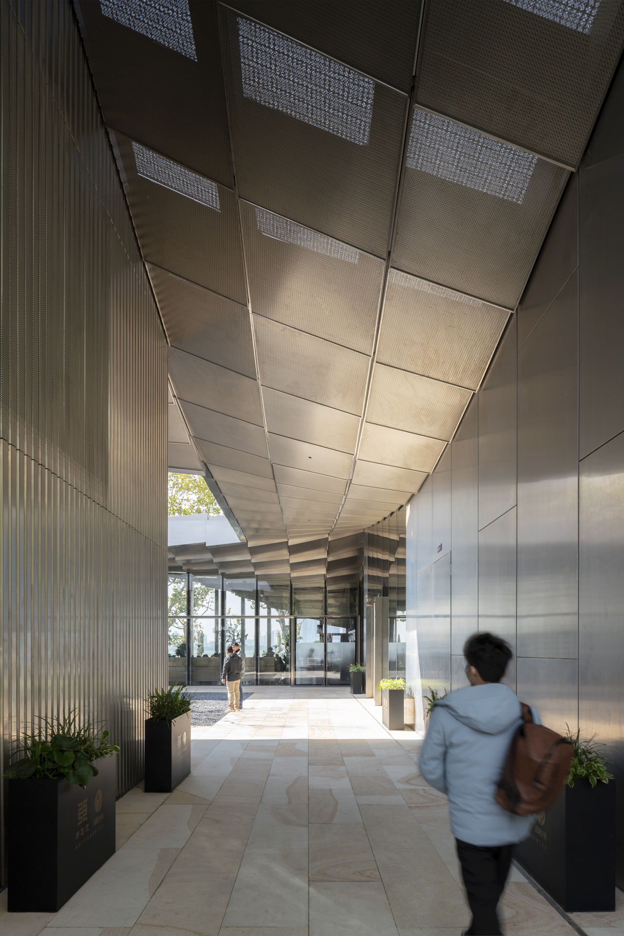 Covered passage with a textured ceiling and planters along the sides. Natural light enters from the courtyard, as people walk through the space.