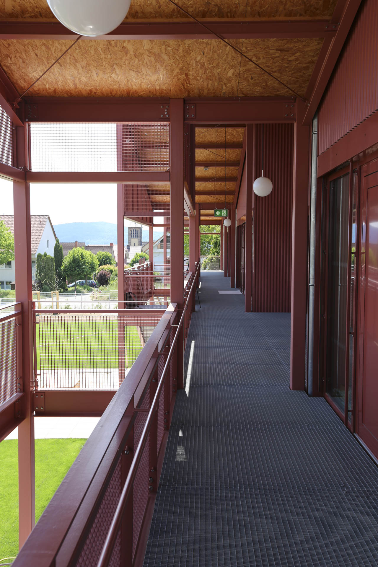 Außenliegender Laubengang im Obergeschoss mit roter Stahlkonstruktion, passend zur gleichfarbigen Fassade.