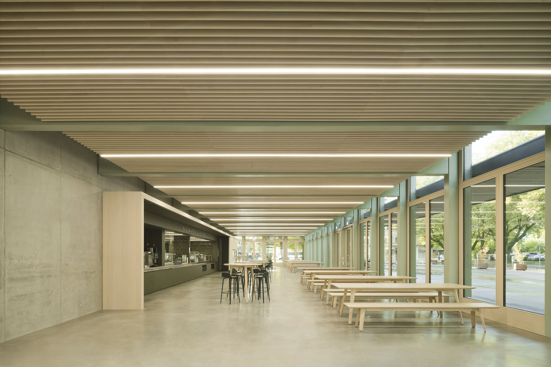Light-filled cafeteria area with high ceilings, warm wooden tables, and fresh green accents.