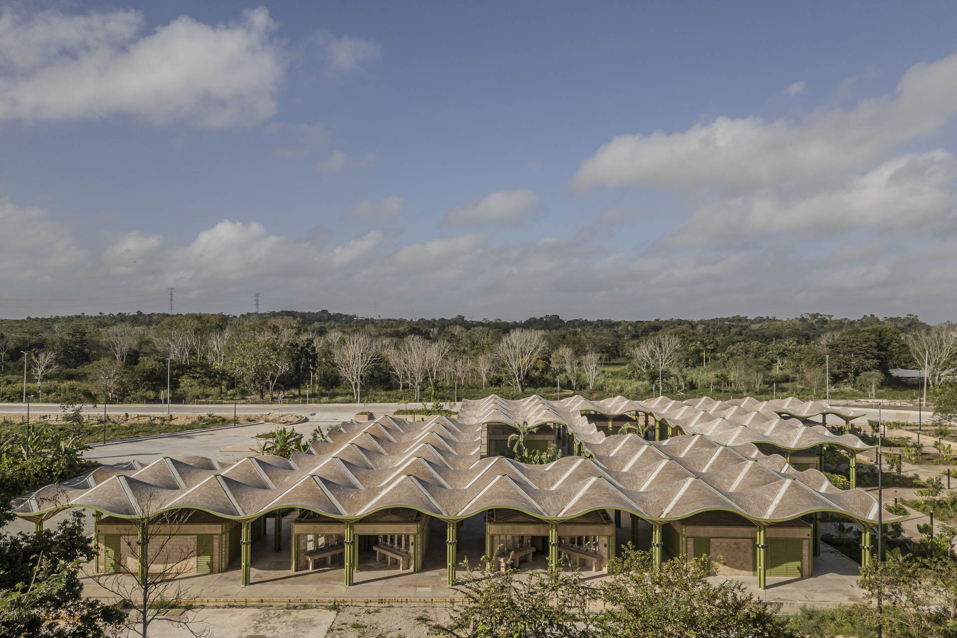 Seitliche Ansicht des Gebäudes mit gewelltem Dach und klarer Rasterung; unter dem Dach Reihen von Baukörpern mit grünen Elementen, umgeben von Sträuchern und Vegetation.