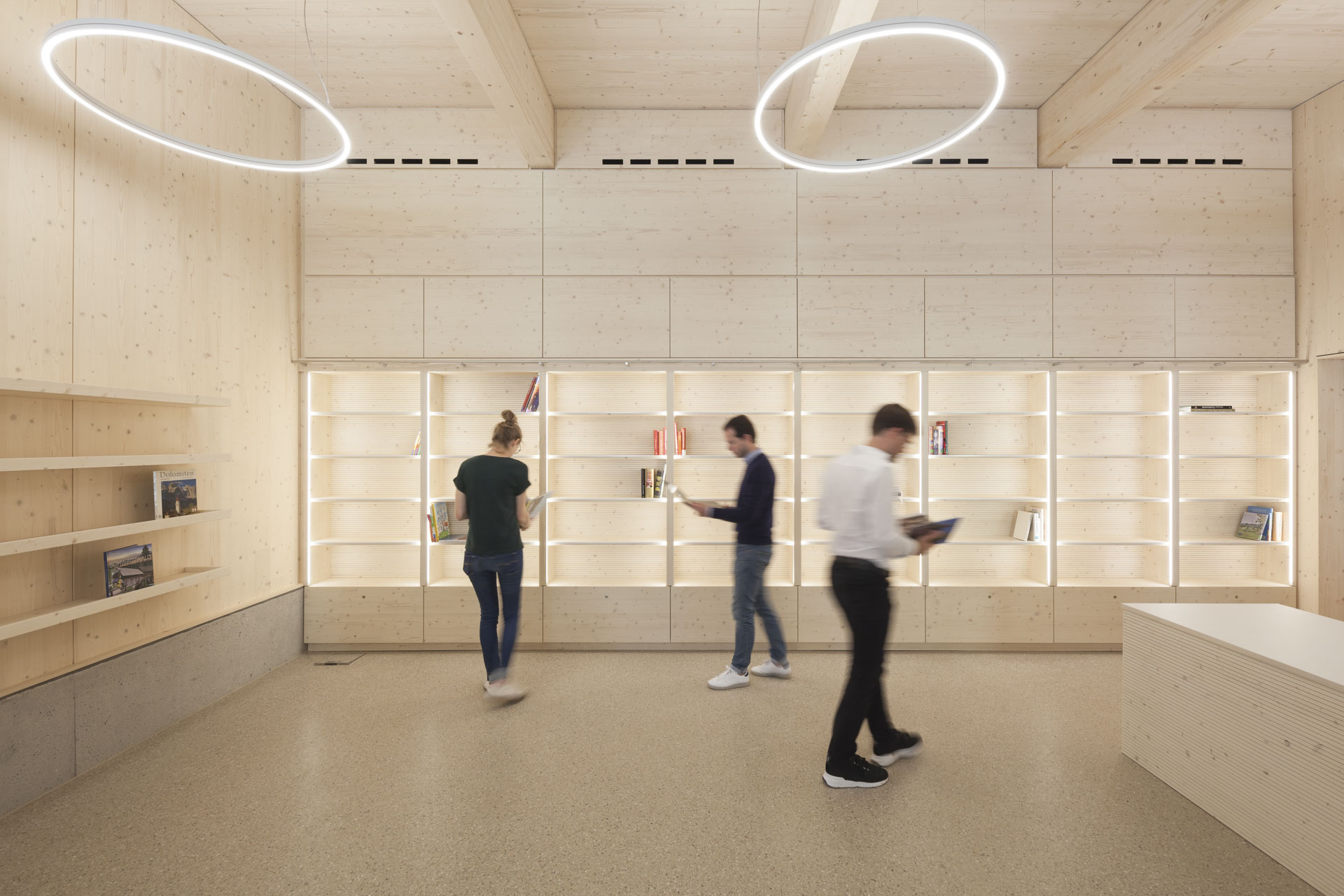 Innenraum mit dominanter Holzausstattung, eingebauten Holzregalen. Personen lesen Bücher im Raum.
