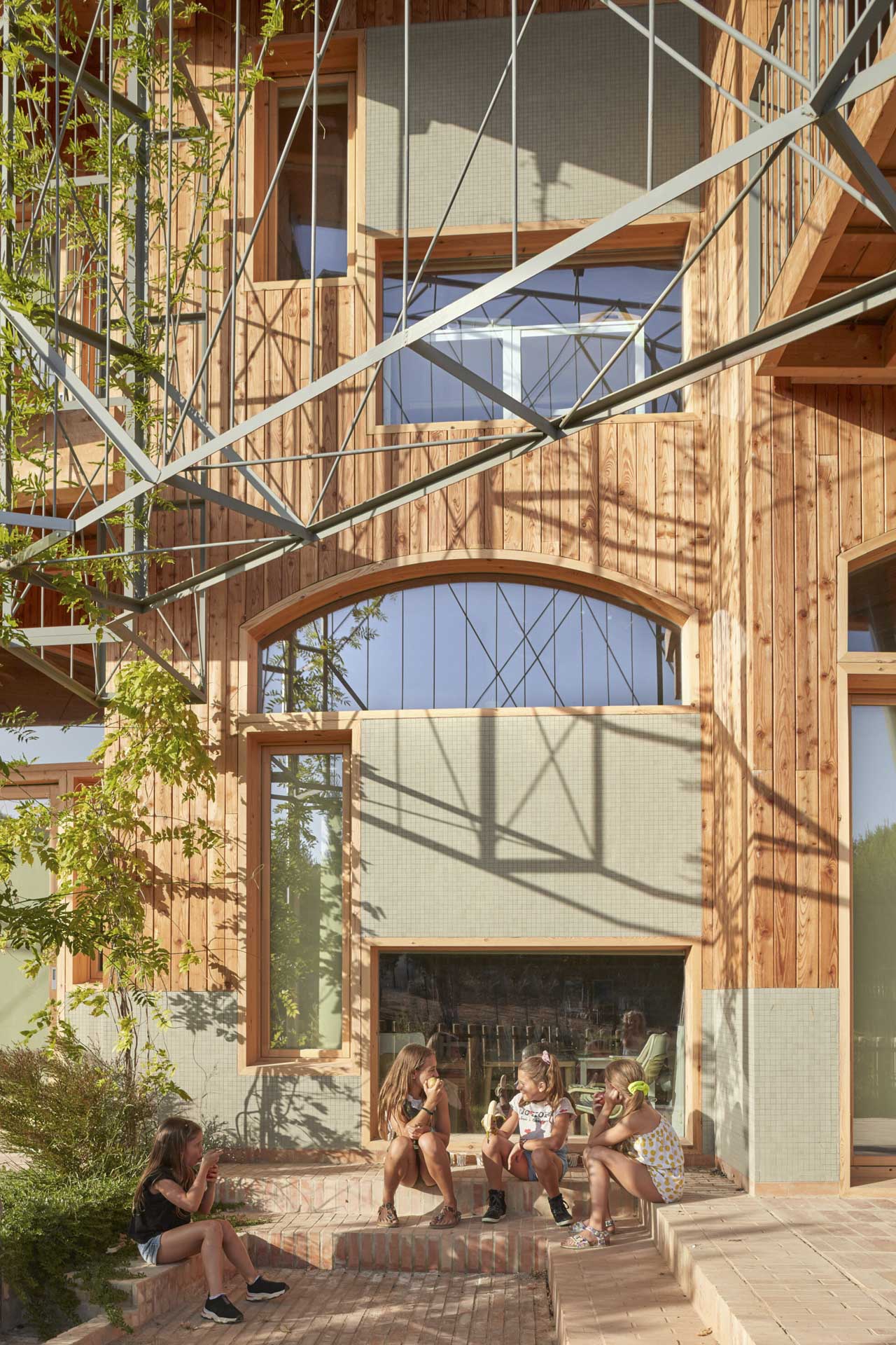 Vier Kinder sitzen im geziegelten Innenhof vor heller Holzfassade mit großen Fenstereinschnitten, umgeben von Bäumen und Begrünung.