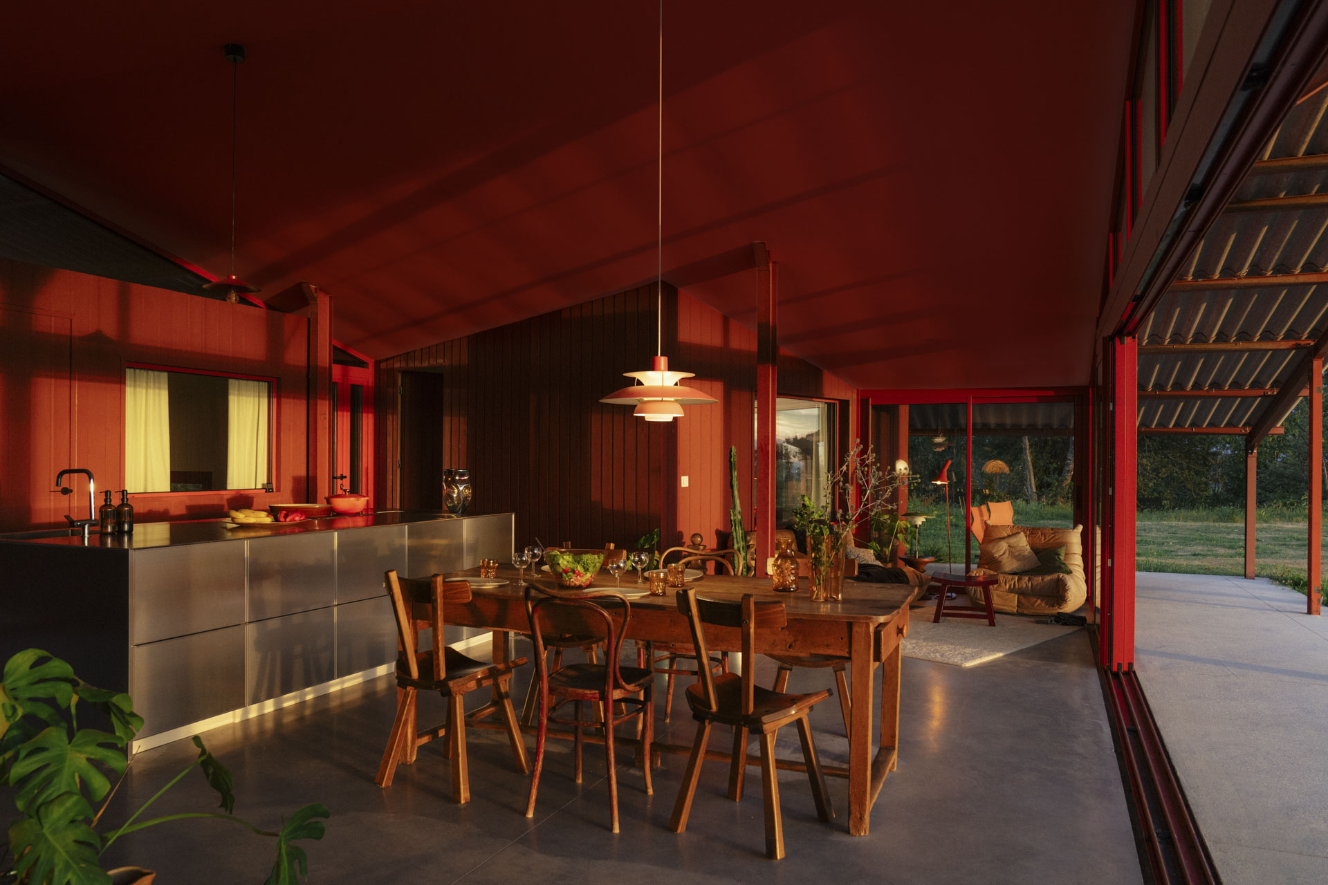 Innenraum offen zur Terrasse und zum Garten, rote Einrichtung, Küche mit Insel, großer Esstisch im Vordergrund.