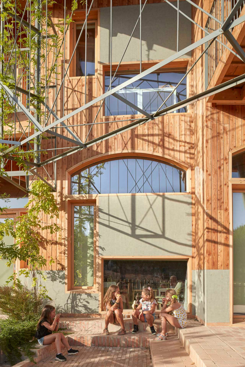 Four children sit in a brick courtyard in front of a light-colored wooden facade with large windows, surrounded by trees and greenery.