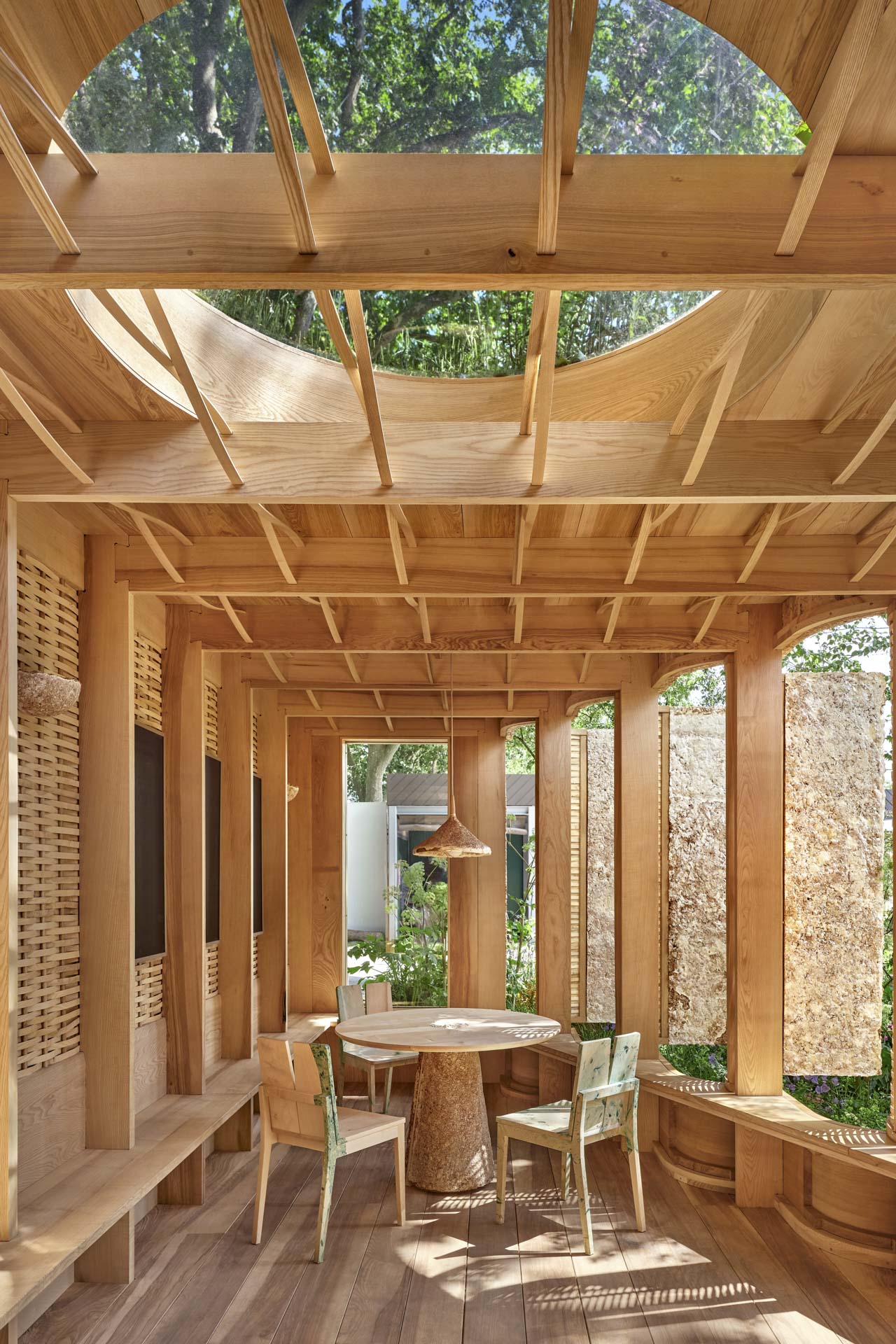 Wohnraum komplett aus Holz mit Tisch und Stuhl, Fenster nach außen geöffnet, durchwachsener Grünraum draußen, rundes Oberlicht belichtet den Raum.