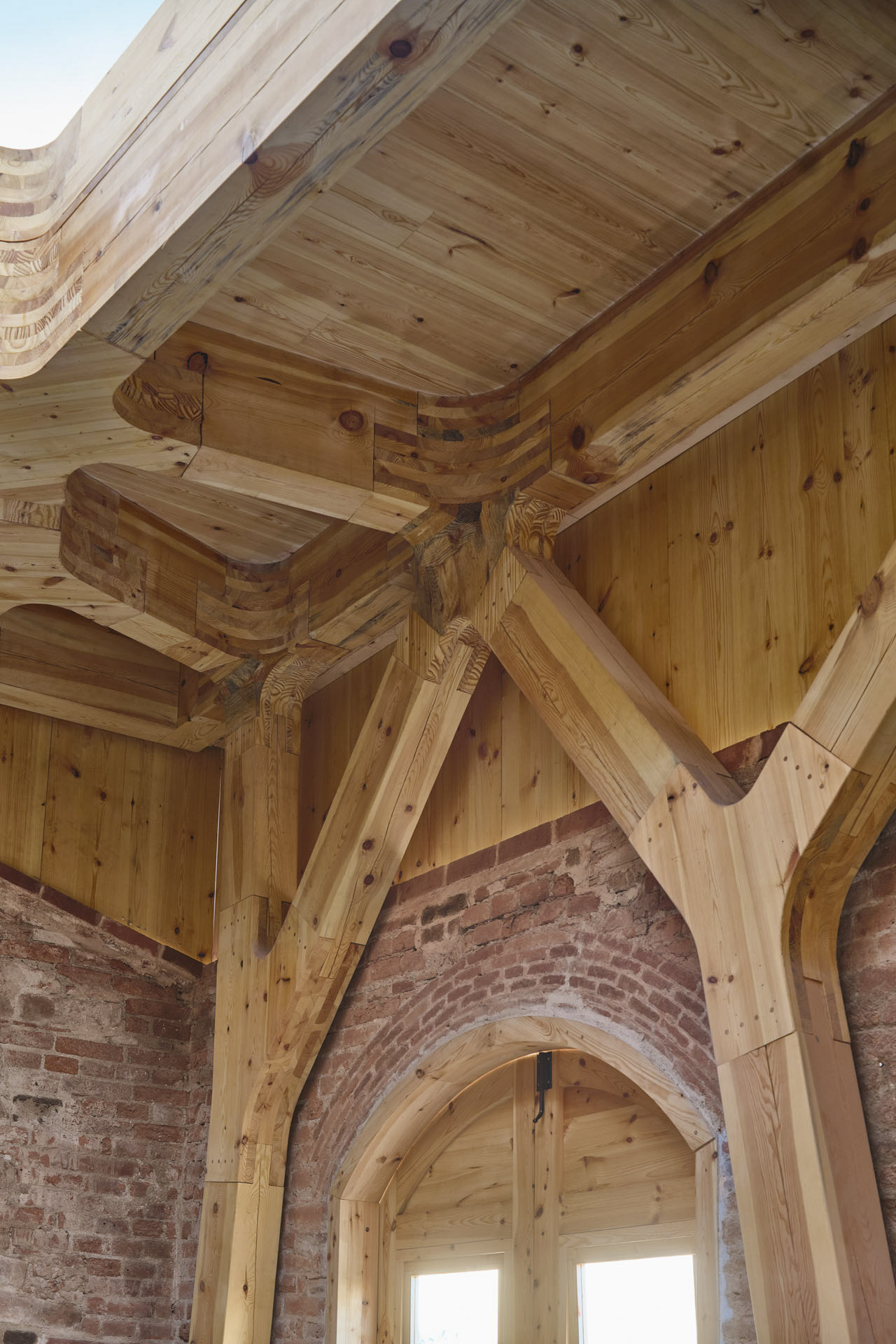 Innenraumecke mit Ziegelwand, Holzdecke mit gefräster Tragwerksstruktur und Oberlicht.