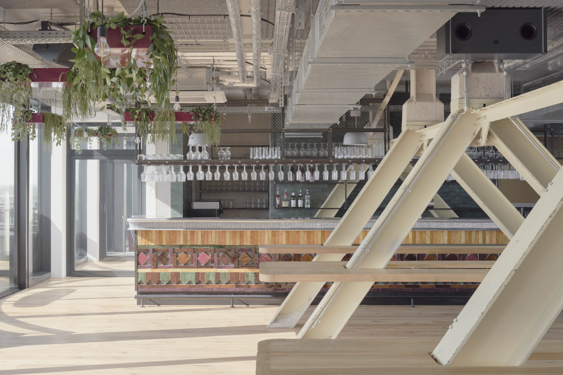 Bar mit farbiger Theke im Dachraum, sichtbare Stahlträger, verglaste Stadtansicht, Betonflächen, offene Schächte und Rohrführungen.