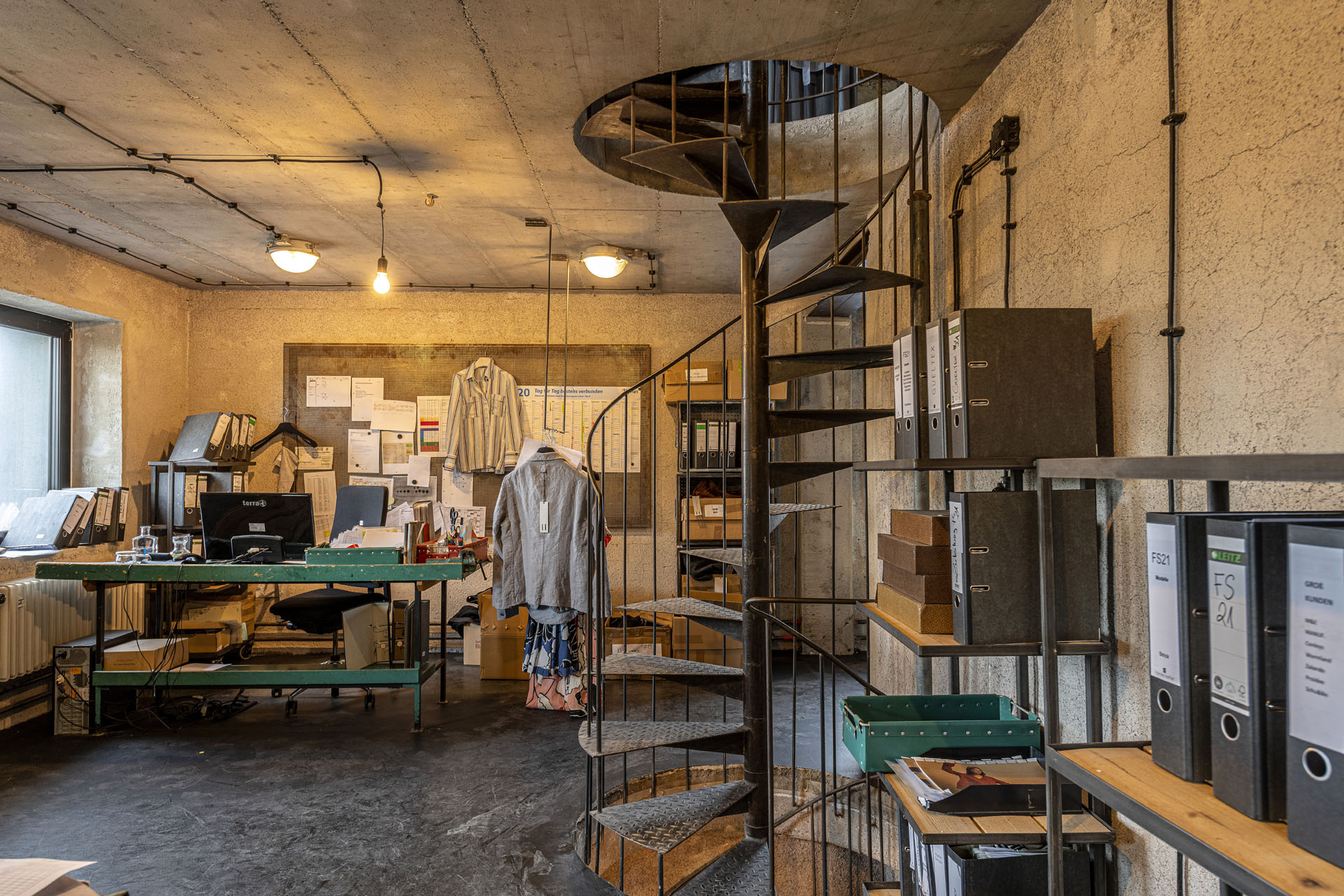 Modeatelier mit stählerner Wendeltreppe und einem Regal mit Aktenordnern rechts und links einem grünen Tisch mit Computer und Büroutensilien.