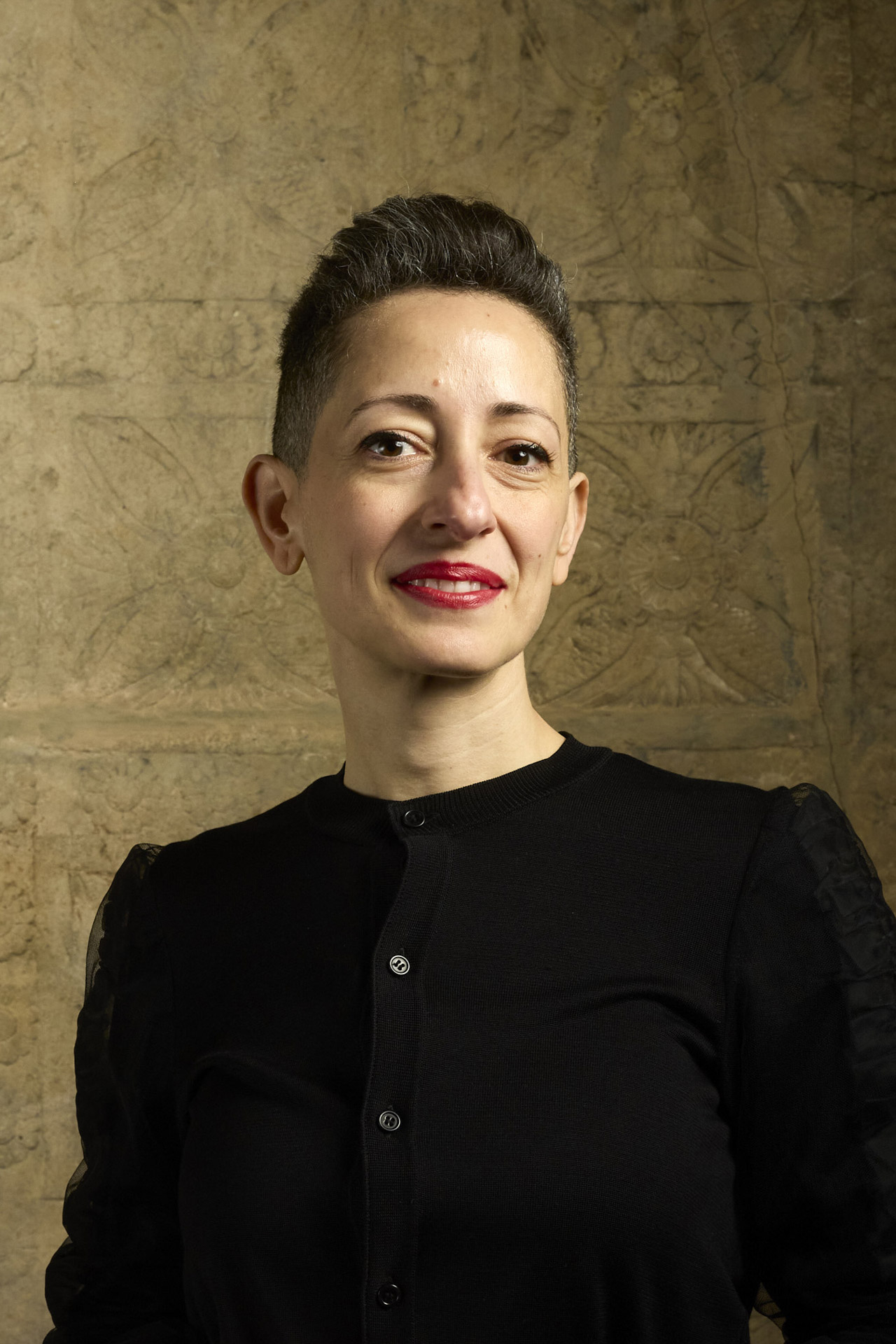 Junge Frau mit kurzem Haarschnitt und roten Lippen, lächelnd, trägt einen schwarzen Cardigan.