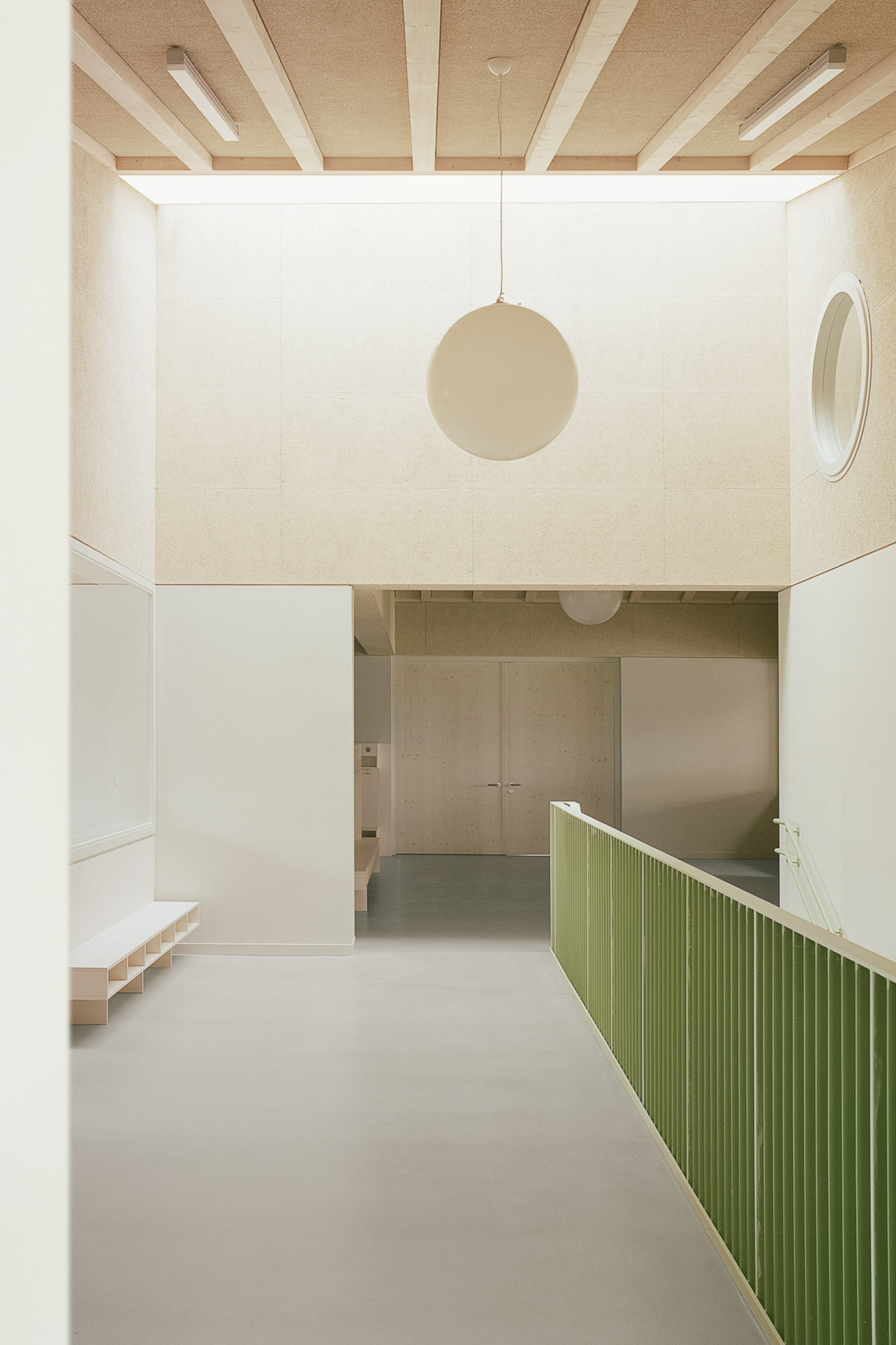 Innenraum mit hellem Holz und hellen Flächen, breiter Gang mit Oberlicht, rechts Treppe mit grünem Geländer nach unten.