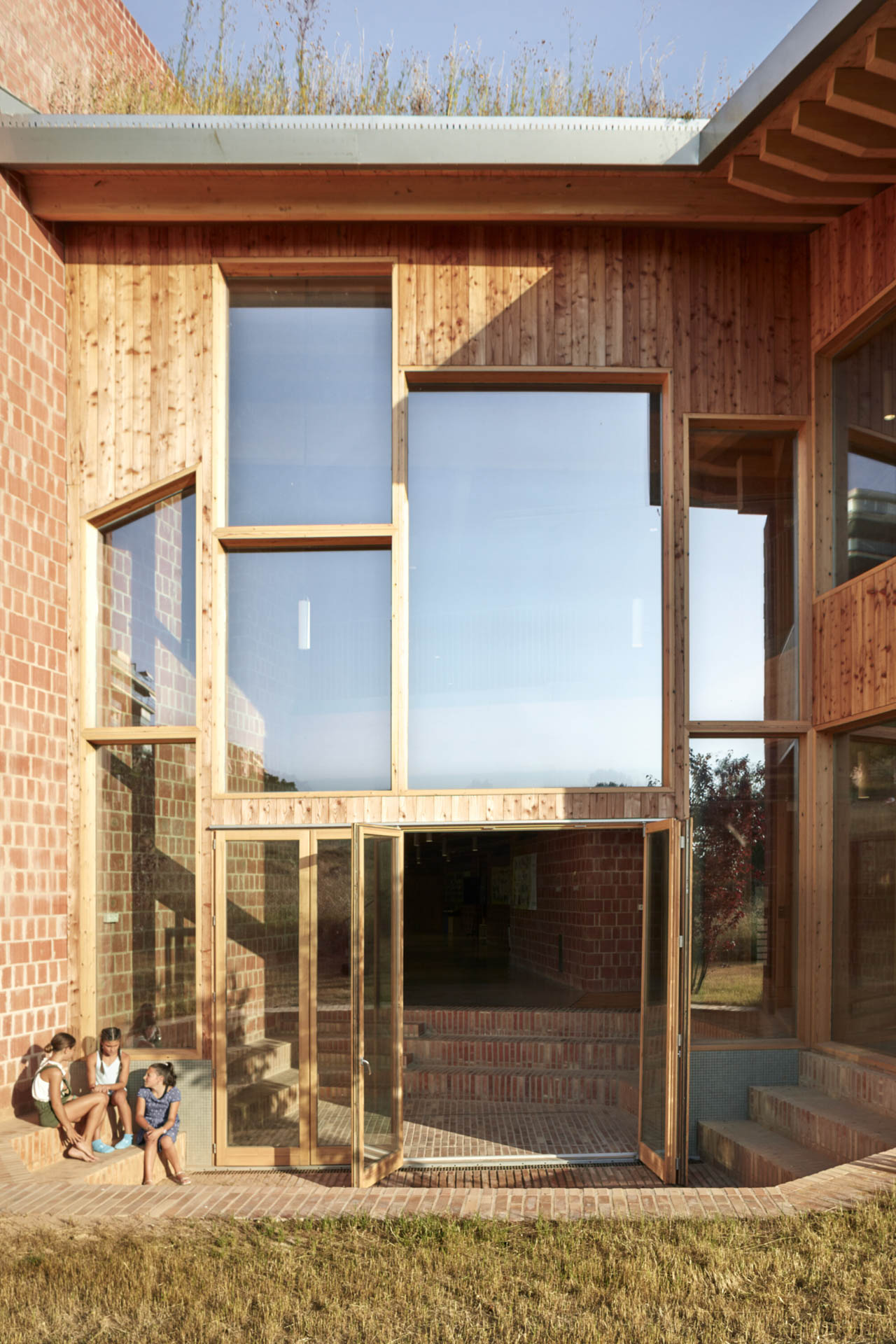 Haus mit Ziegel-Holz-Fassade und variierenden Fenstern, geziegelte, bodenversenkte Terrasse. Kinder sitzen und unterhalten sich.