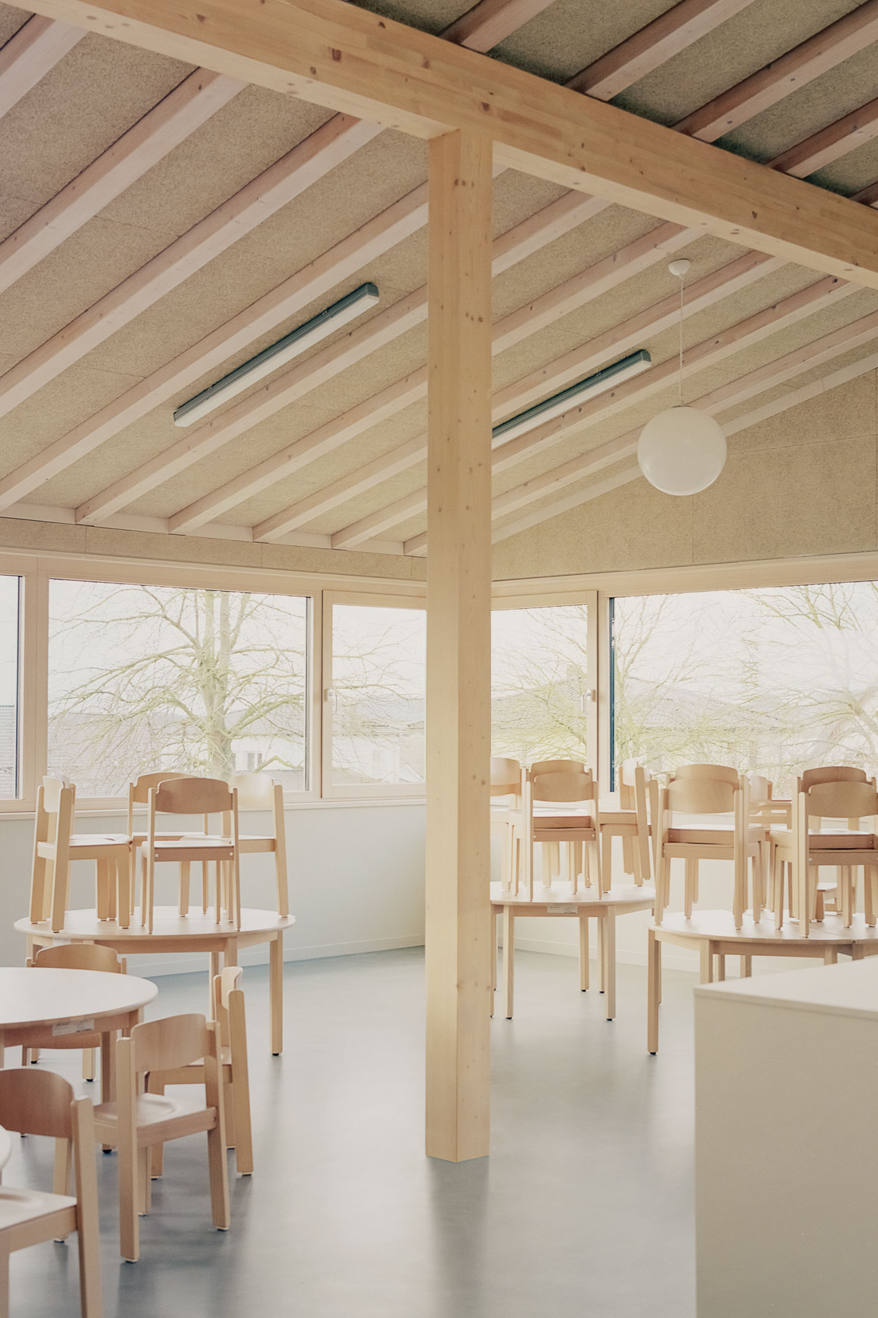 Innenraum mit runden Holztischen, je vier Holzstühle verteilt im Kinderessraum, horizontale Fenstergliederung.
