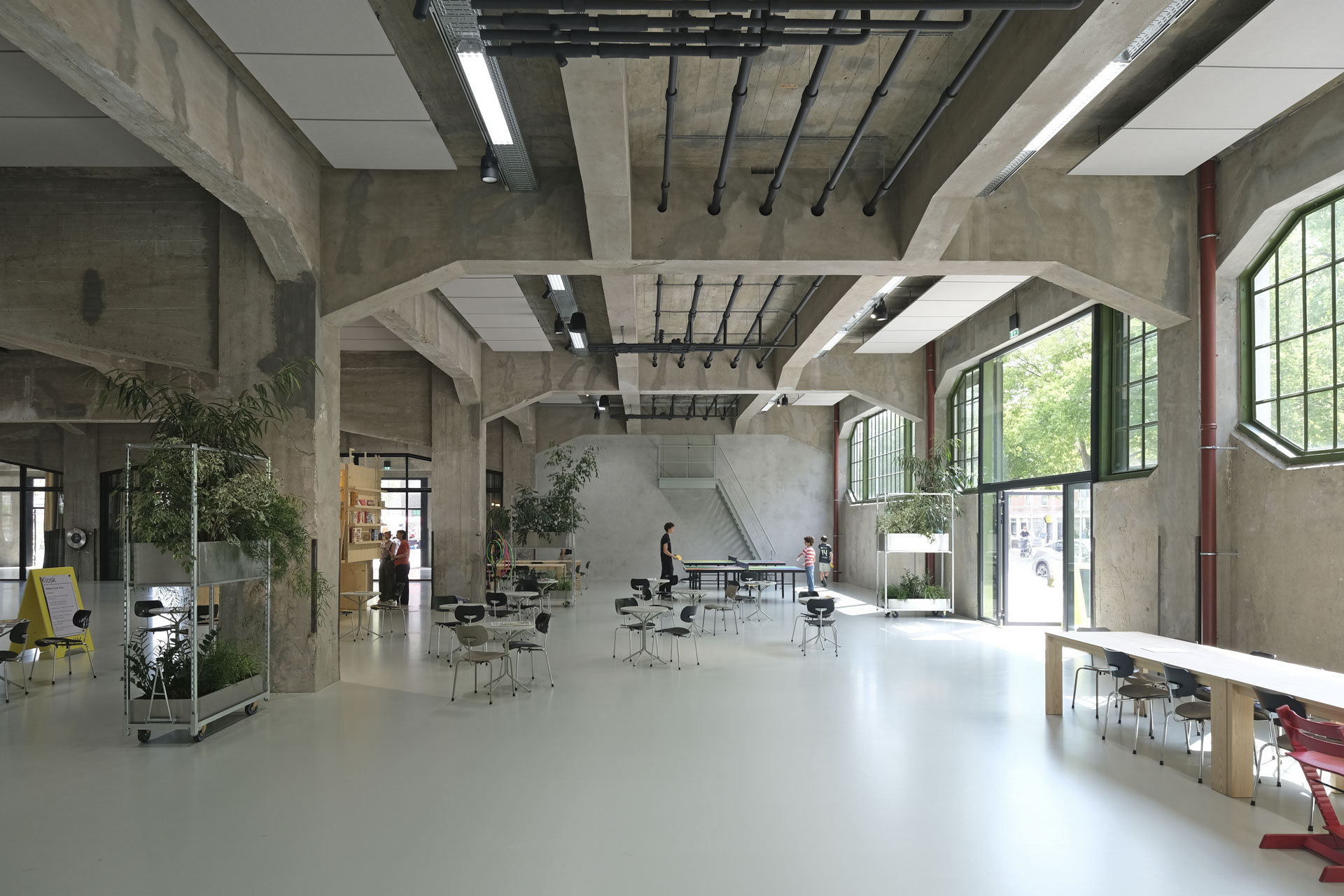 Concrete-dominated interior with high ceilings, massive columns and beams, large green-framed factory windows illuminate the hall.
