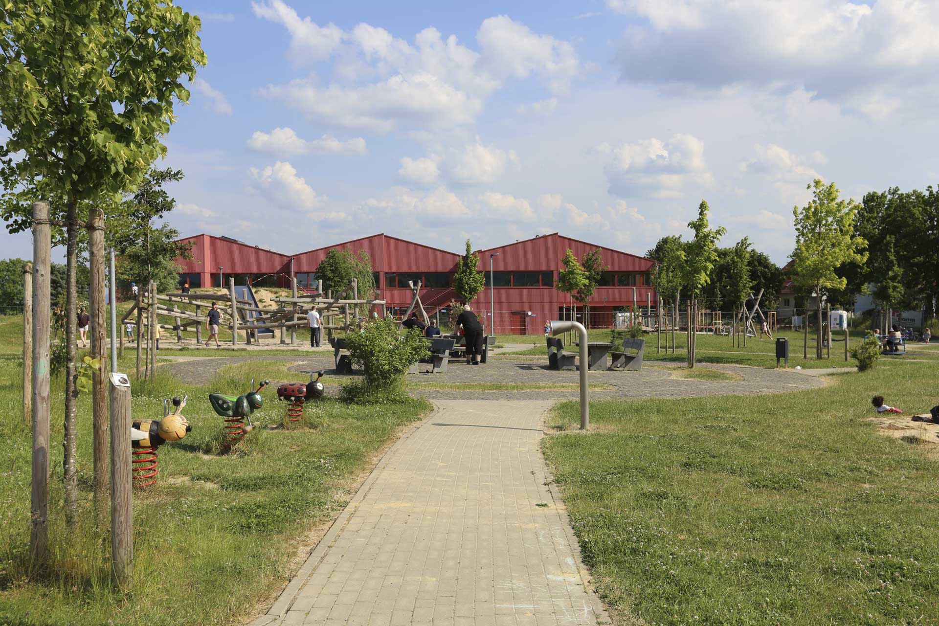 Haus mit drei Satteldächern und roter Metallfassade in grüner Landschaft mit Spielplatz.