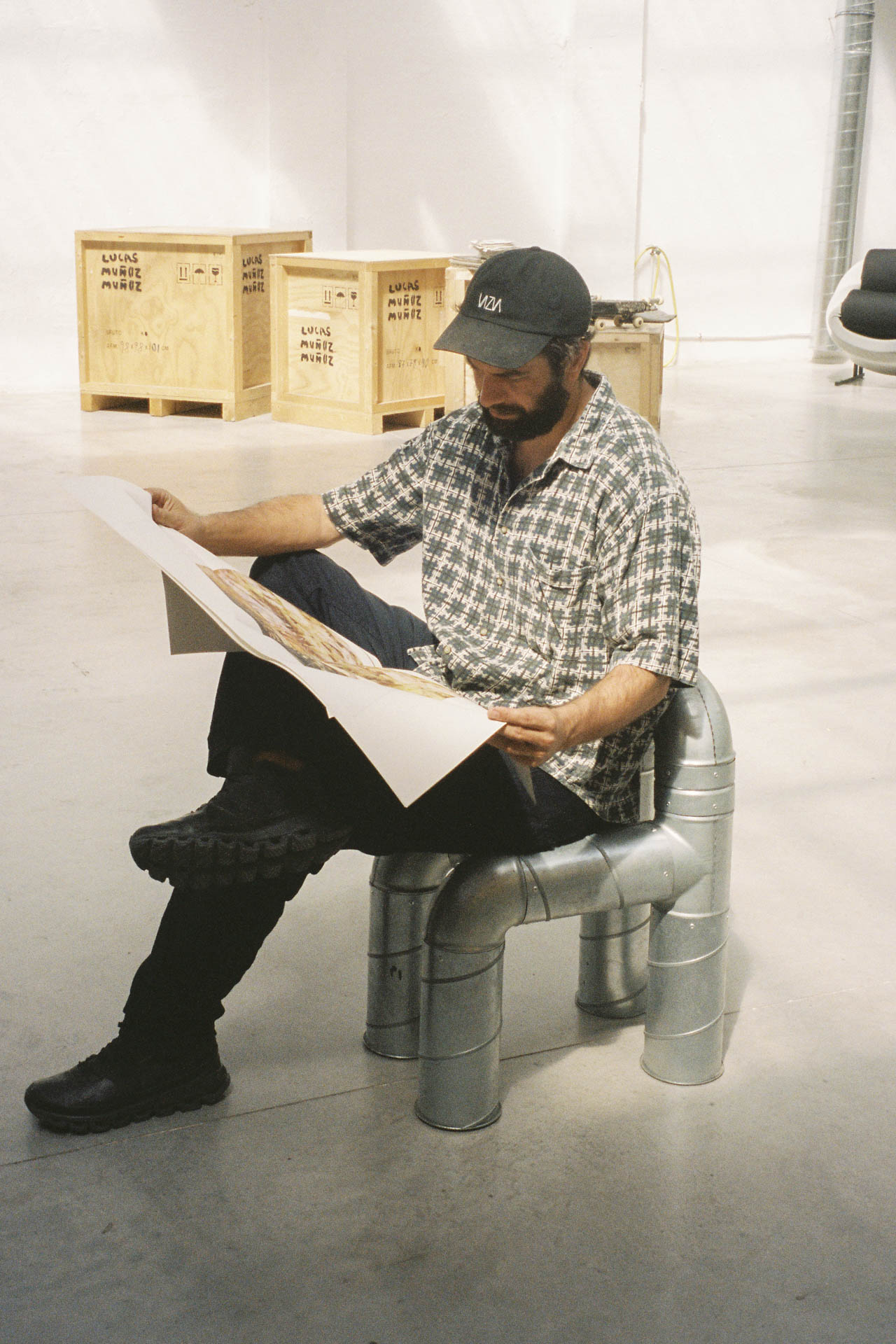 Junger Mann mit Bart und Cappy in lässigem Hemd sitzt auf einem Metallstuhl, hält eine große aufgeklappte Zeichnung; im Hintergrund gestapelte Kisten mit seinem Namen.