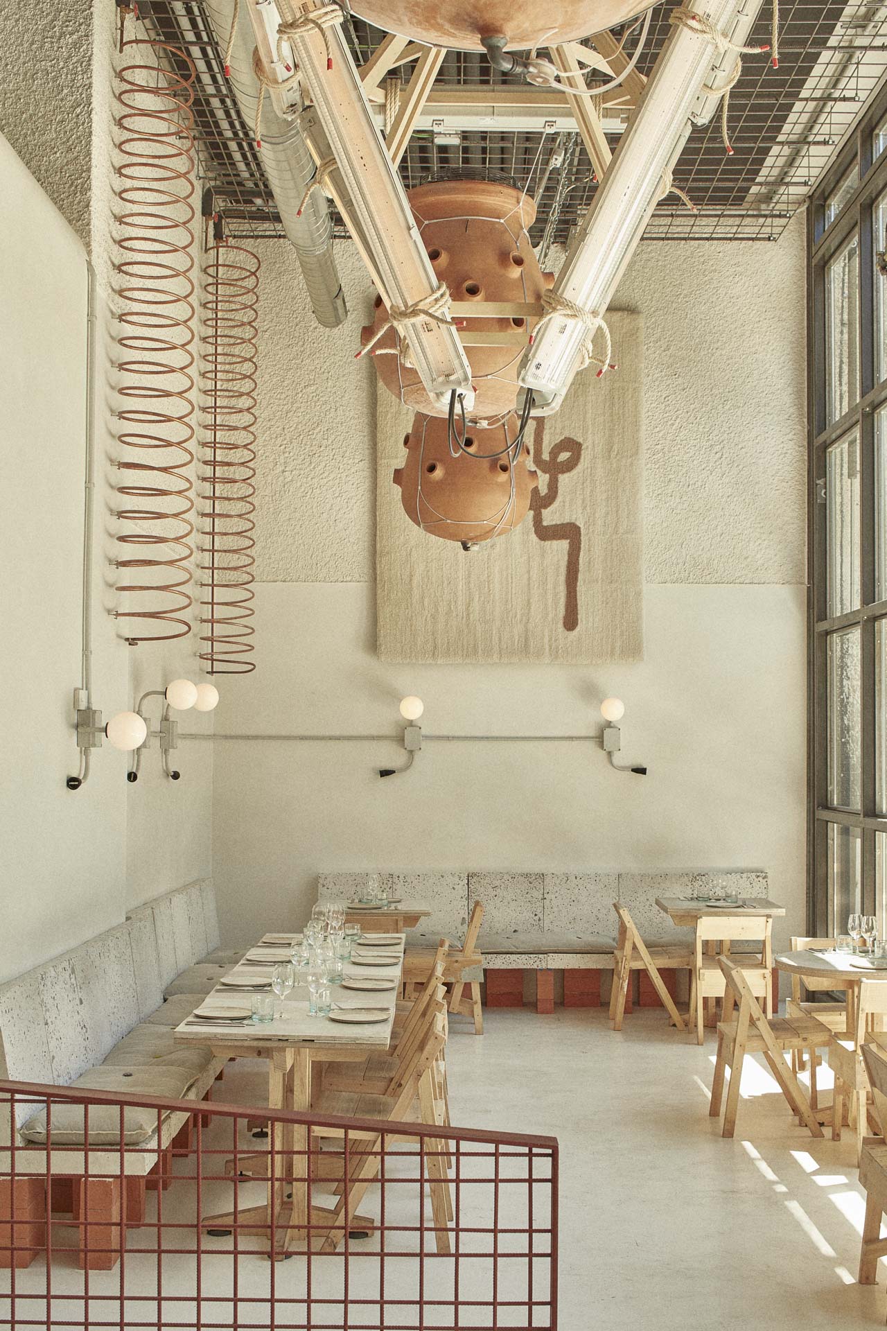 Heller Raum mit hohen Wänden, sichtbarer Technikdecke, minimalistischen Holzbänken und Wandlampen, zentrale Installation aus Metall und Keramik.