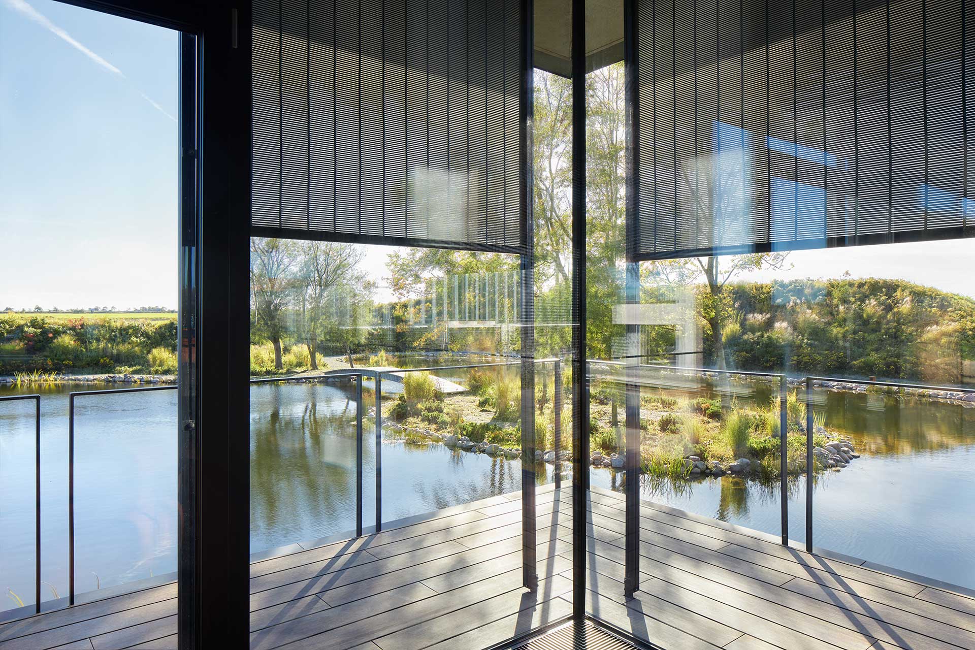 Blick durch ein Glaseck auf den au&szlig;en liegenden See mit gr&uuml;nem Ufer.