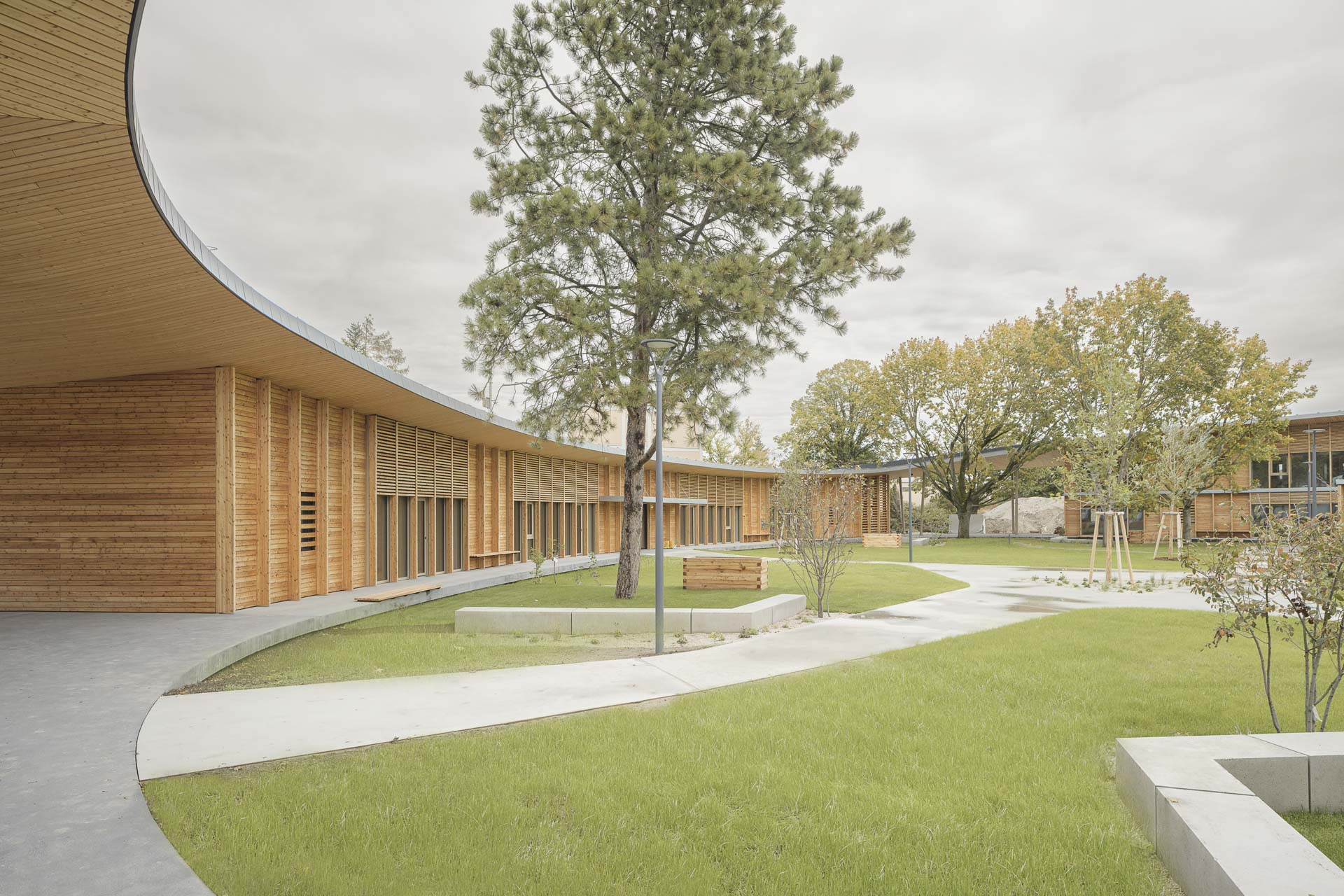Flacher, geschwungener Holzbau mit vertikaler Lamellenfassade, großen Fensteröffnungen und vorgelagertem Grünraum mit Wegen und Bäumen.