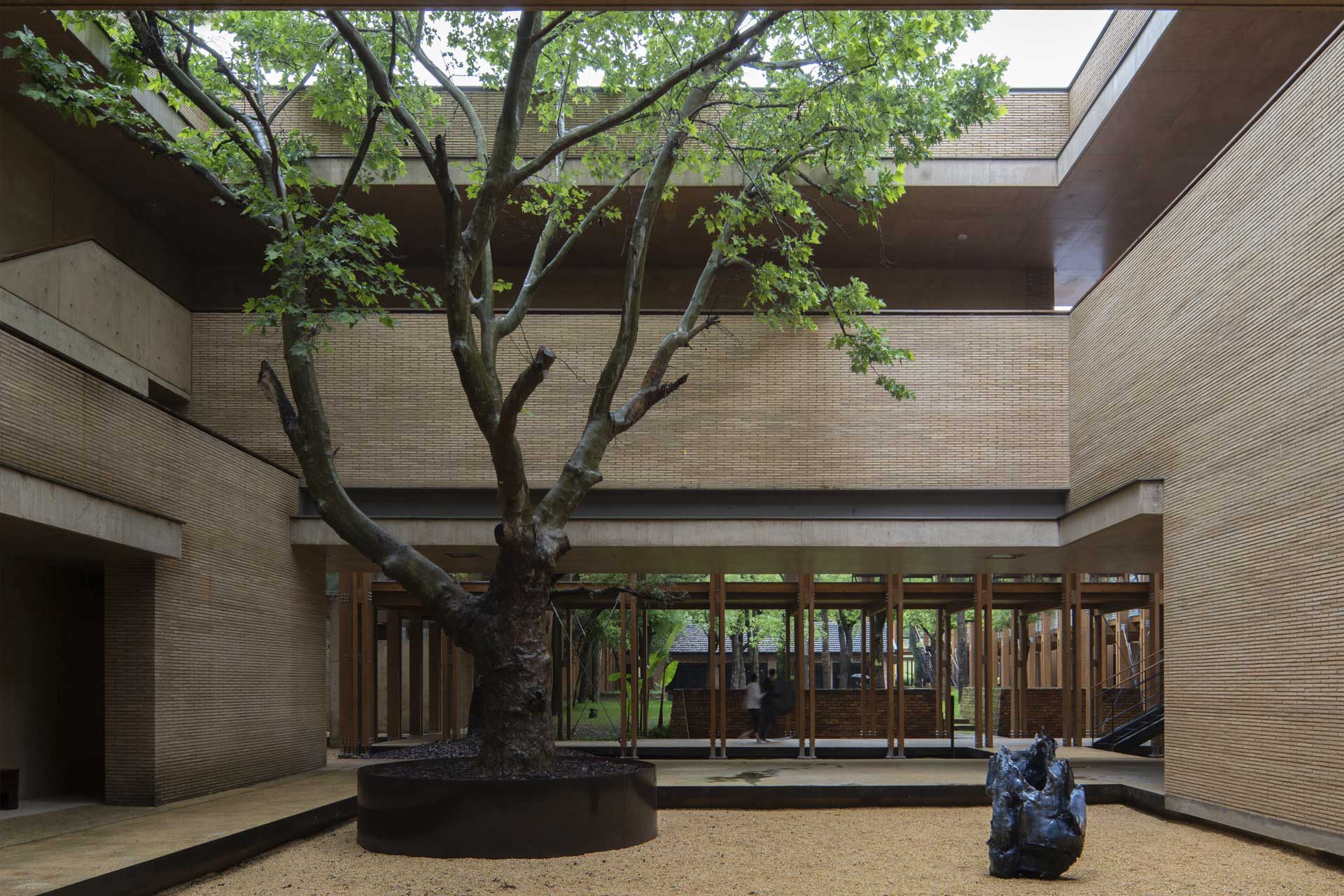 Innenhof mit großem Baum in zweigeschossigem Gebäude, geschlemmte Ziegelfassade, Fensterreihe mit offener Glas-Holzfassade im Erdgeschoss.