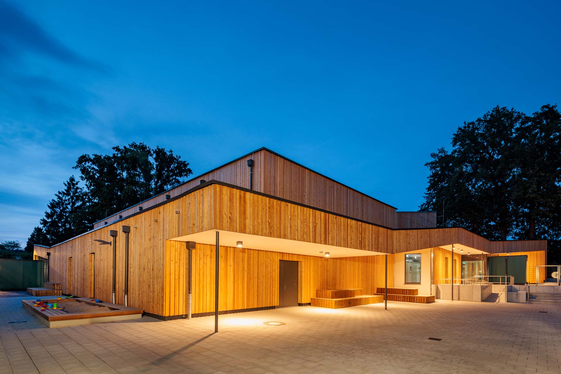 Beleuchtetes Holzgeb&auml;ude mit Flachdach und Laubengang.