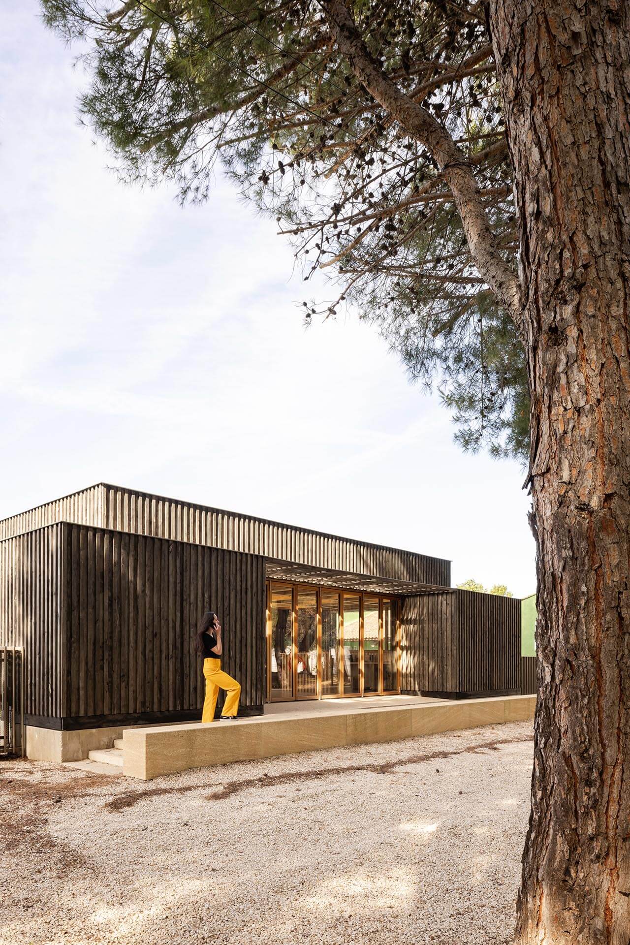 Bungalow mit Holzverkleidung. Auf der Terrasse steht eine Frau mit gelber Hose.