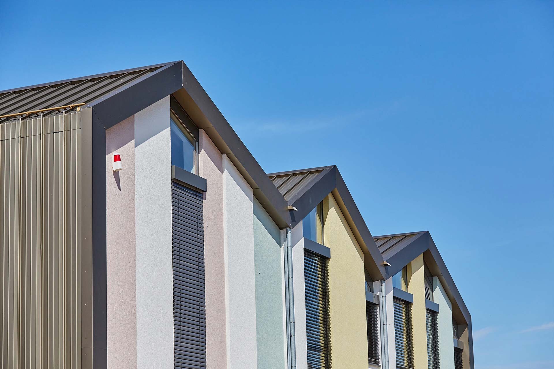 Obere Teile eines Geb&auml;udes mit drei A-f&ouml;rmigen Dachspitzen und bunten Streifen an der Hauswand.