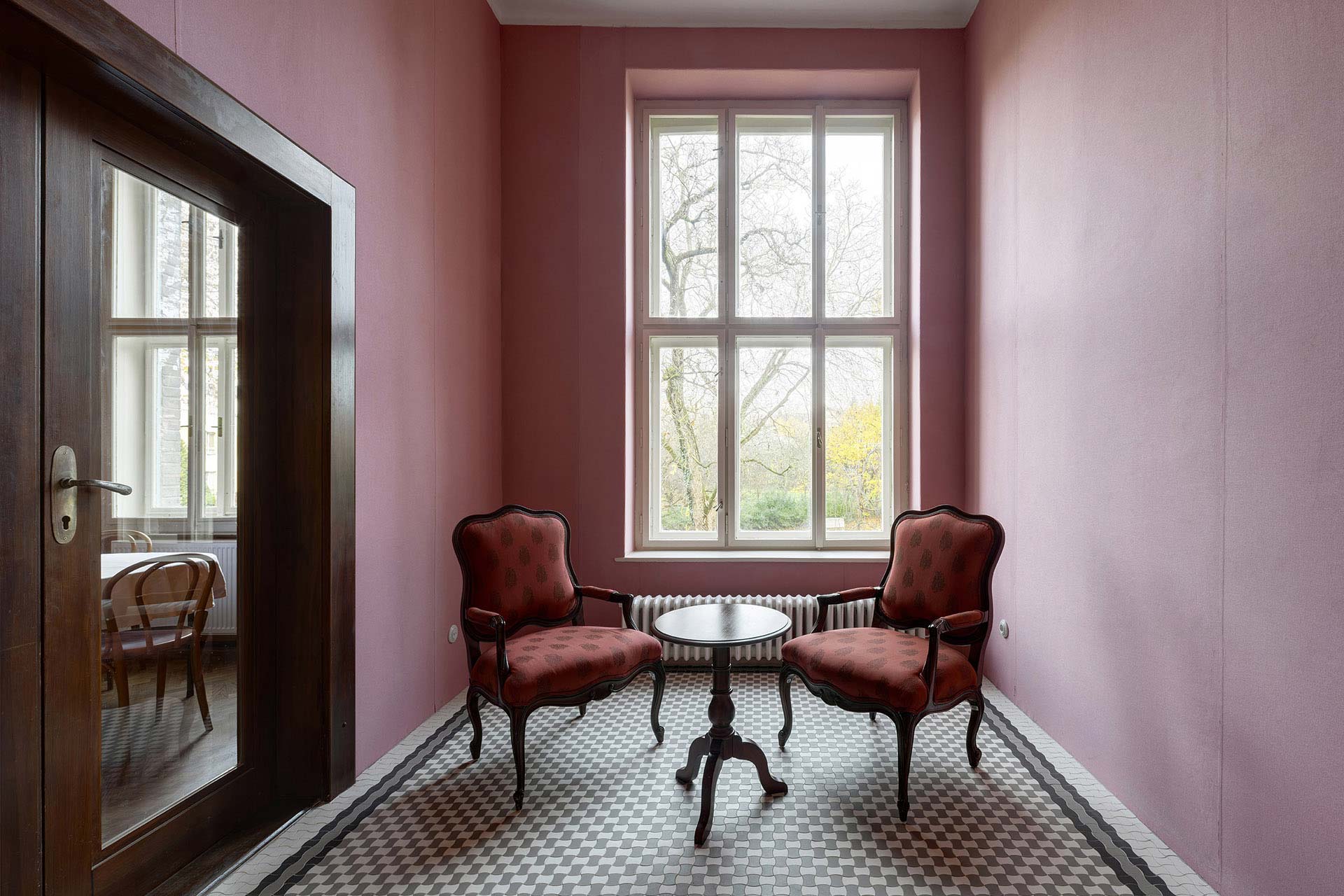 Leerer, herrschaftlicher Raum mit Fischgr&auml;tparkett, dunkel get&auml;felten W&auml;nden und einer verzierten Decke. Die bodentiefen Fenster &ouml;ffnen den Blick in den Garten.