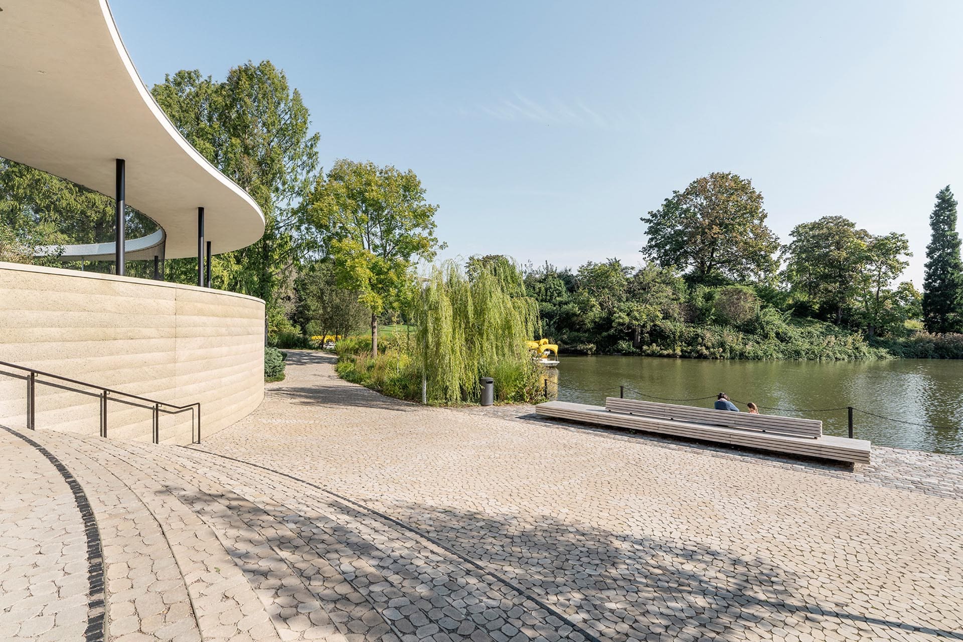 Gepflasterter Bereich am See mit Treppen und Sitzgelegenheit.