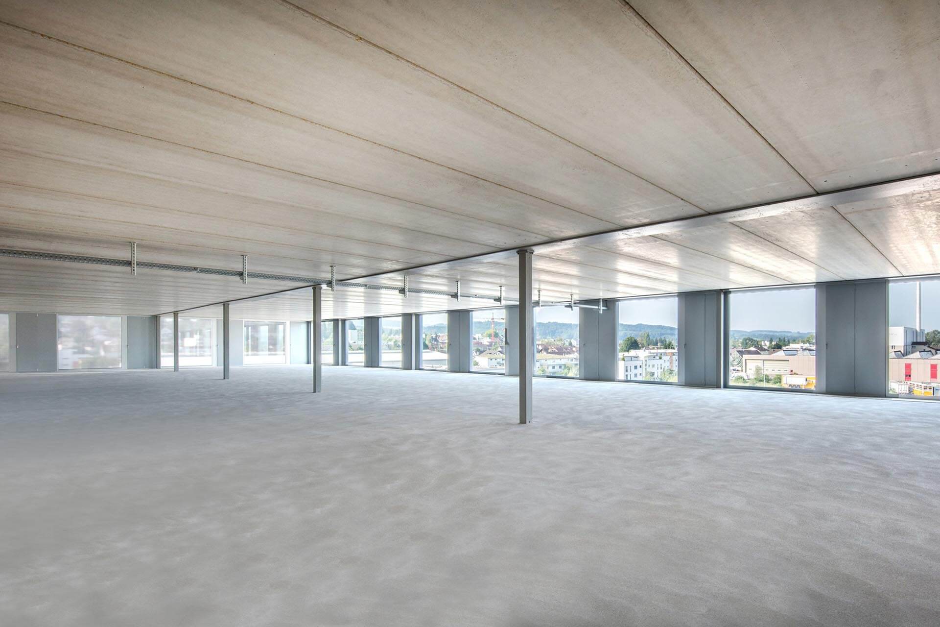 Großer, leerer Raum mit vier schlanken Stützen mitten im Raum. Durch die bodentiefen Fenster schaut man auf eine Stadt.