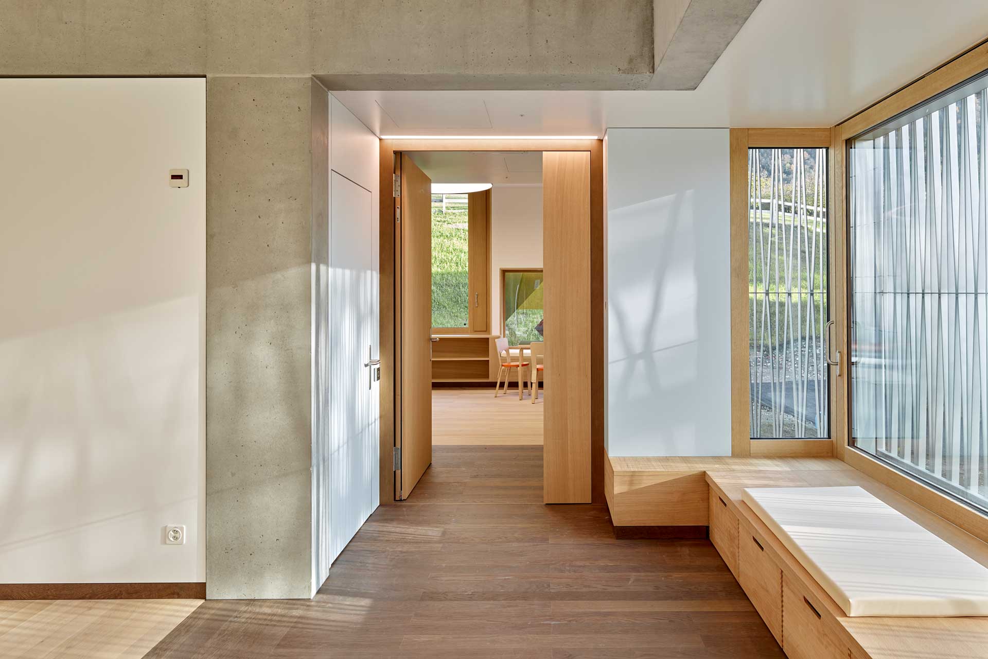 Flur mit PArkett und Sitzfläche vor dem Fenster. Hinten steht eine Tür offen, man blickt in ein Patientenzimmer.