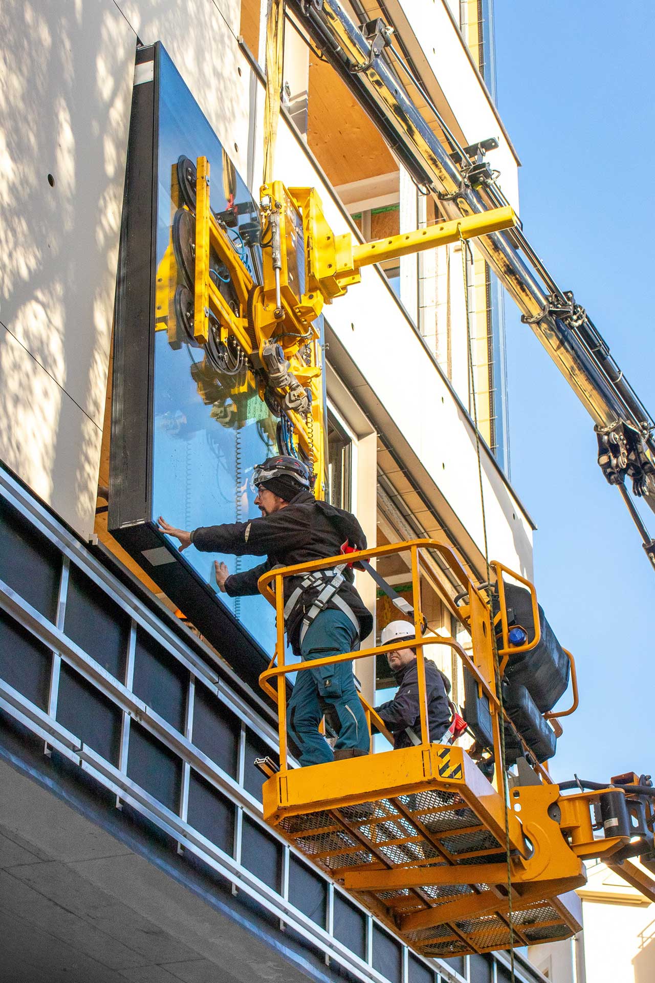 Zwei Arbeiter auf einem Hebekran montieren ein Fenster an einem Gebäude.