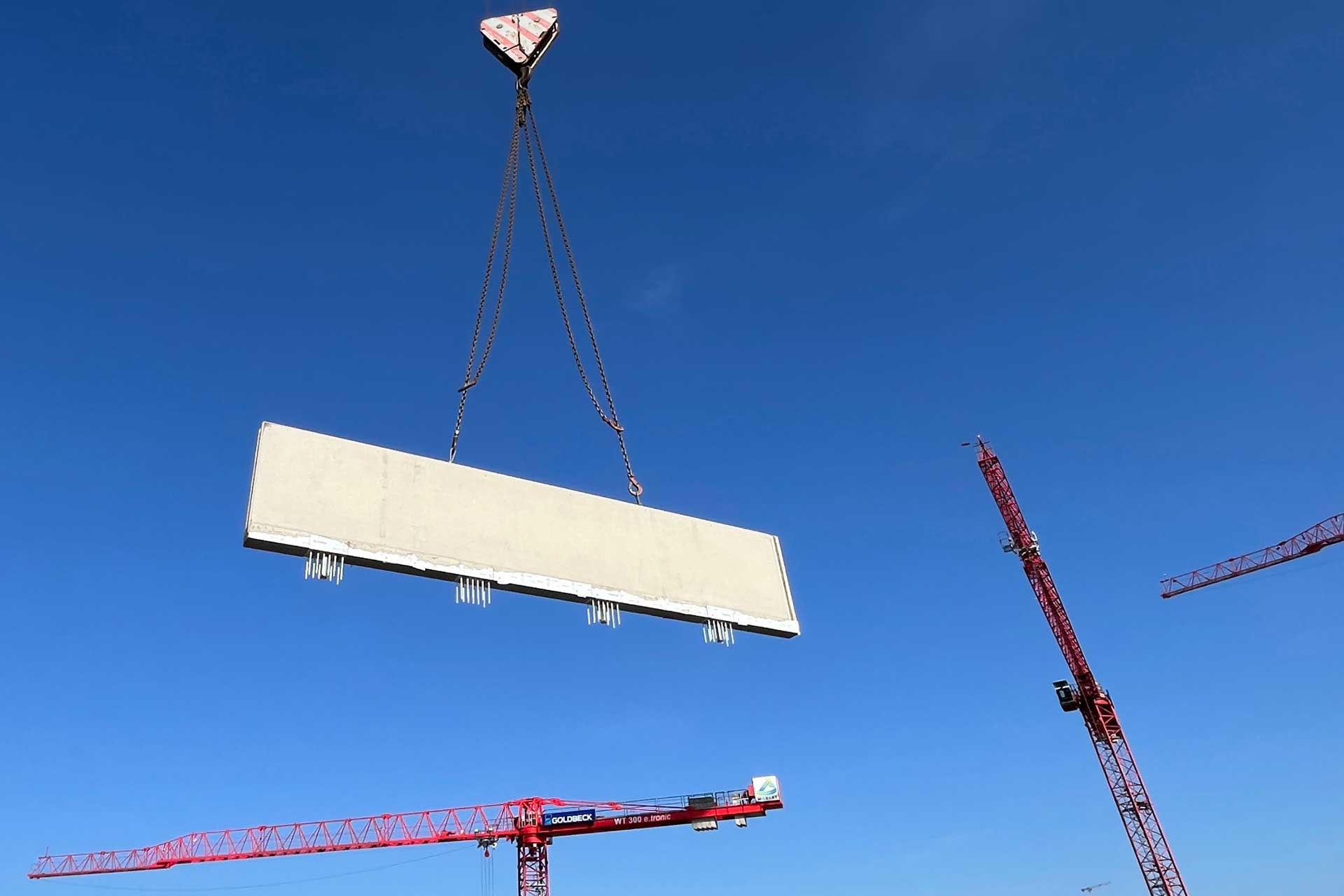 Ein Betonfertigteil hängt an einem Kran in der Luft vor blauem Himmel.