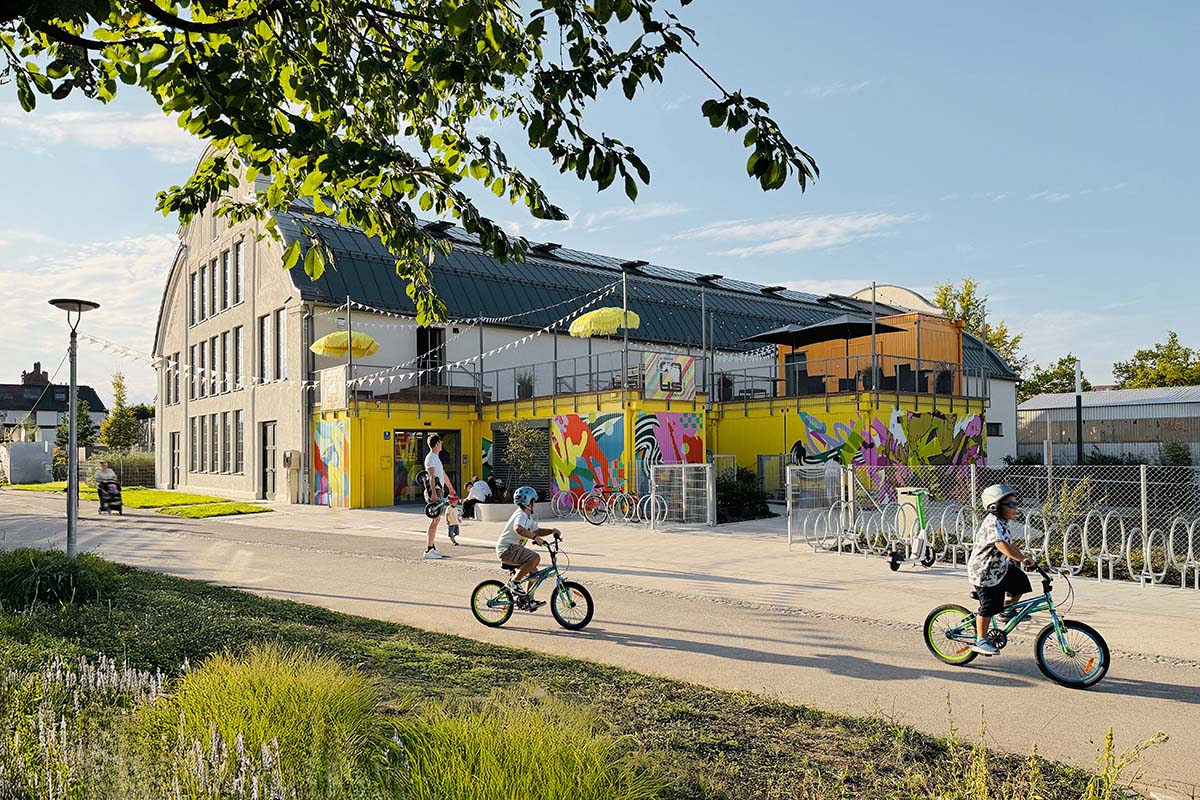 Kinder fahren Fahrrad im Vordergrund; im Hintergrund Altbau mit saniertem Dach, gelber Containeranbau mit bemalten Wänden und Dachterrassen.