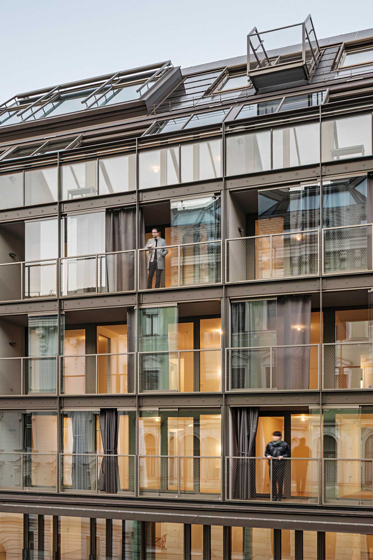 Hausfassade mit verglasten Balkonen und drei Menschen, die auf verschiedenen Balkonen stehen.